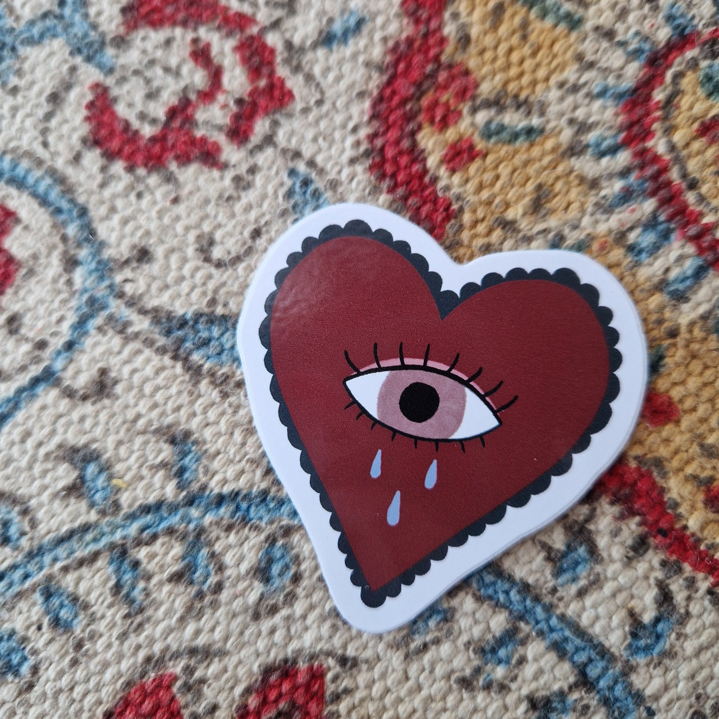 Closer view of the red heart sticker with dark blue frills around the edge and a pale pink crying eye in the middle. Behind the sticker is a patterned fabric in white, yellow, blue and red.
