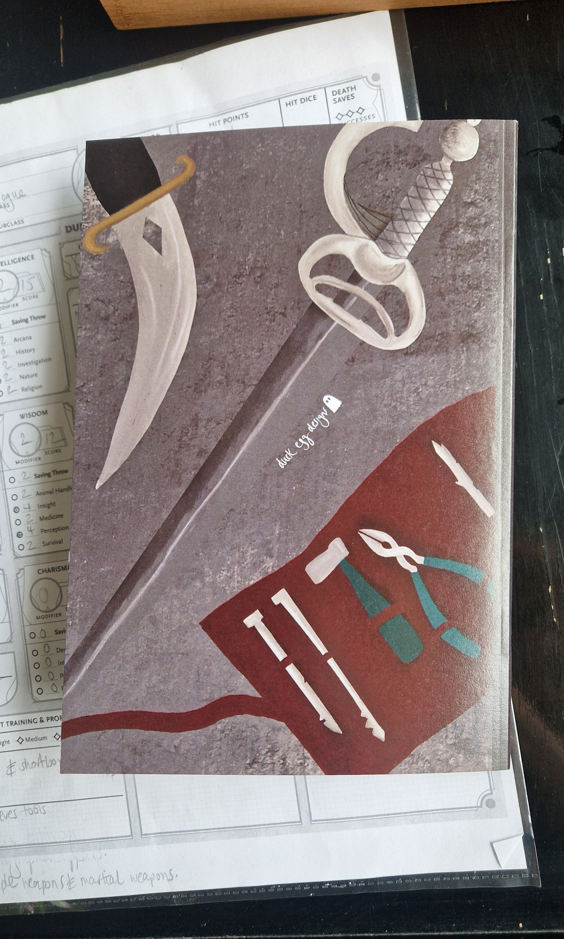 Back of the cunning actions notebooks showing a dagger and sword as well as a thieves tool pouch. The notebook is grey and the tools pouch is a deep red. A small duck egg designs logo is on the back in white.