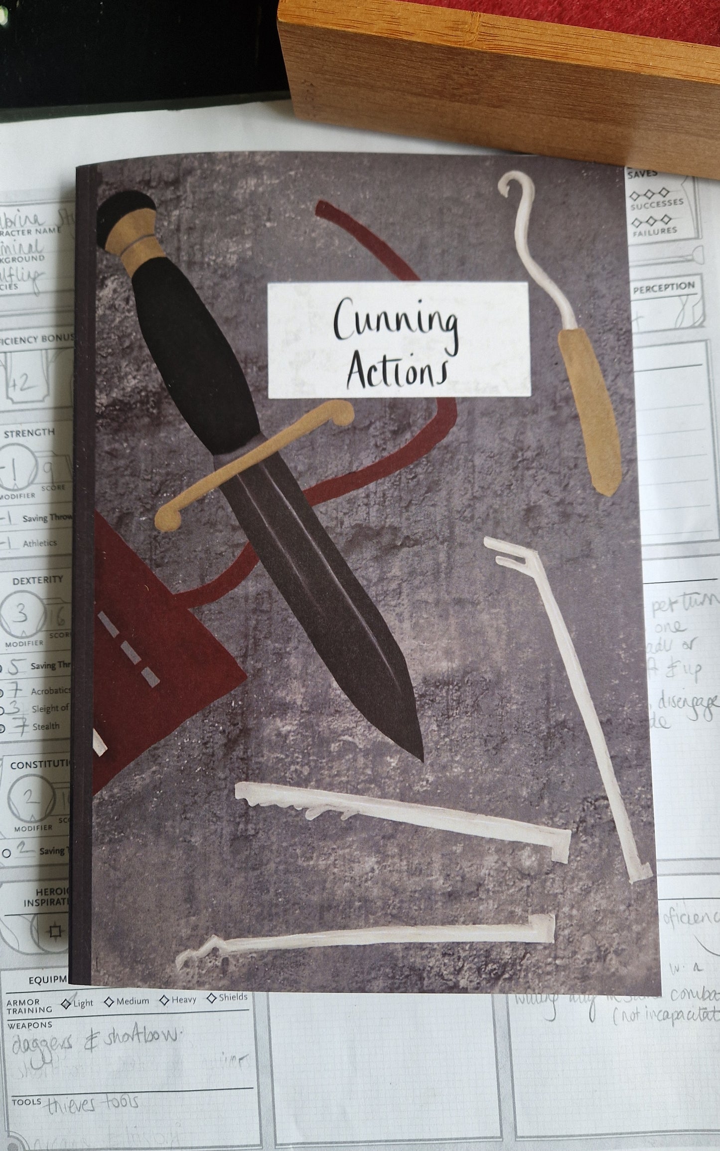 An A5 grey notebook featuring a dagger and thieves tools and the words 'Cunning Actions' in black handwriting in a white box. Behind the notebooks is a character sheet and you can see a dice tray just above it.