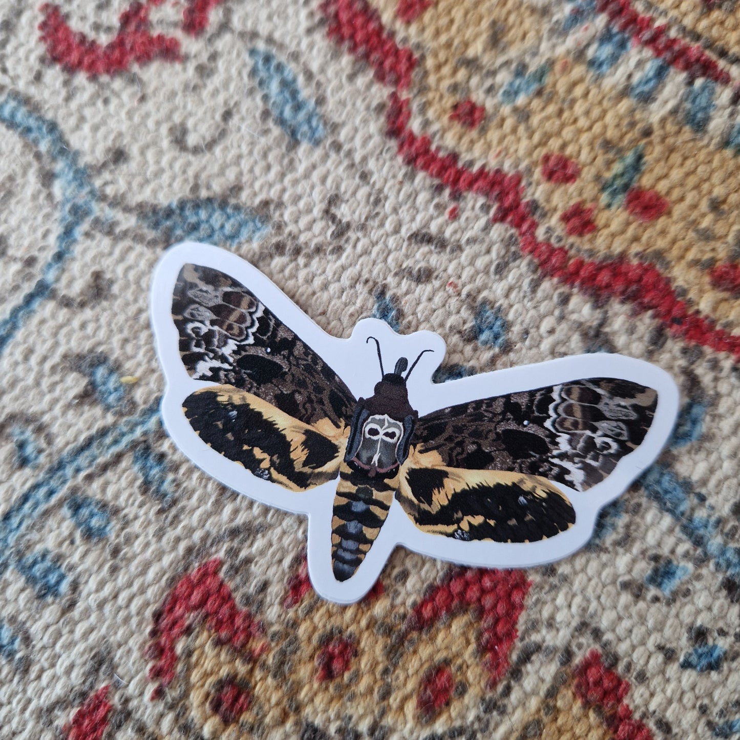 Orange, brown, white, grey and black death head moth sticker on a cream, red yellow and blue patterned fabric. The moth has a skull like motif on its back.
