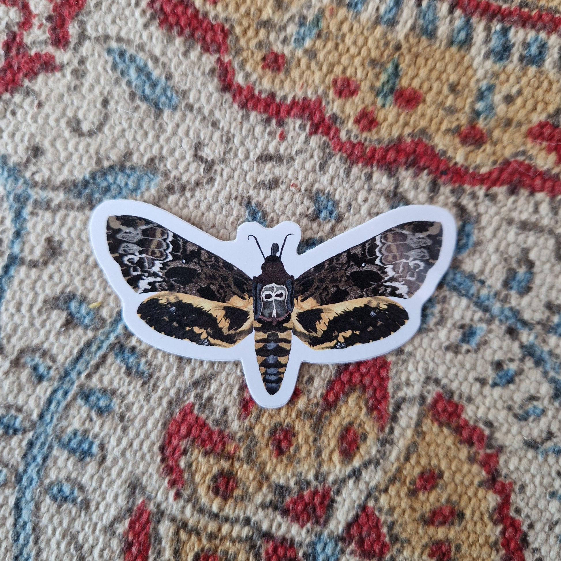 Death head moth sticker on a cream, yellow, blue and red patterned fabric. The moth is brown, orange, white, grey and black with a skull like motif on it's back.