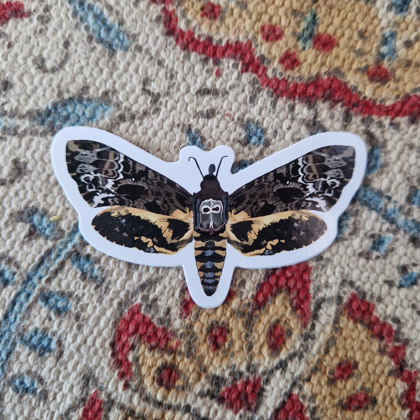 Orange, brown, white, grey and black death head moth sticker on a cream, red yellow and blue patterned fabric.