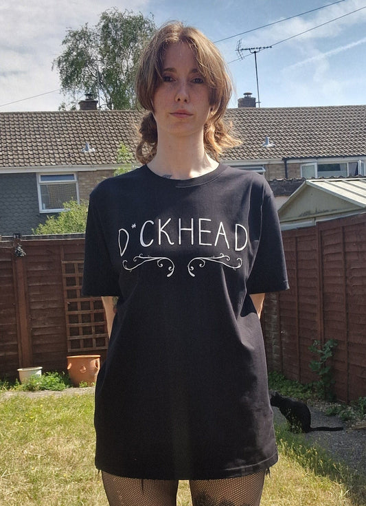 A white woman wears a black t-shirt with the word 'D*ckhead' written across the chest with a filigree inspired design running underneath the writing. Behind her is a green garden and a small black cat.