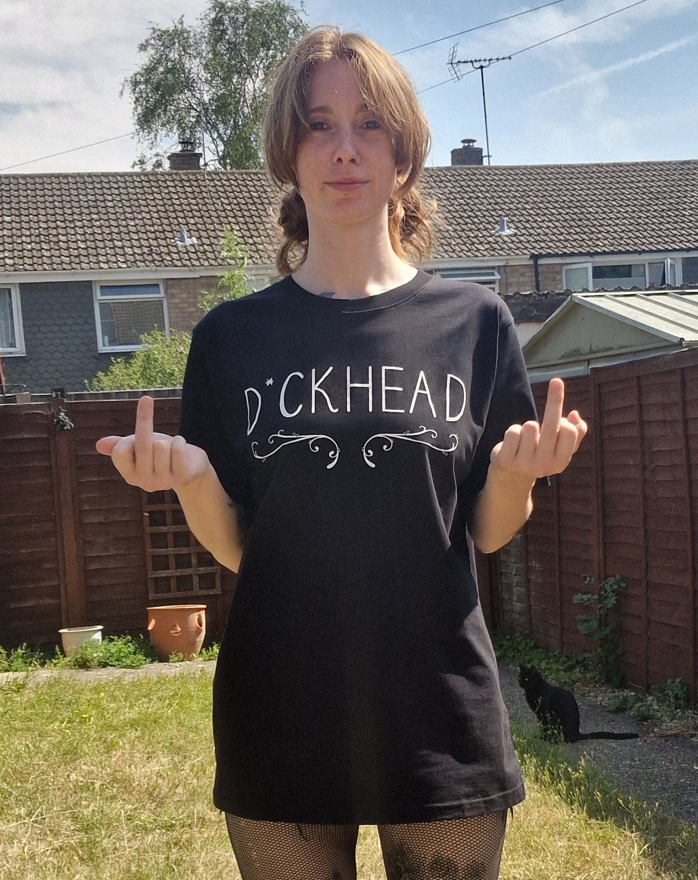 A white woman flips the bird while wearing a black t-shirt with the word 'D*ckhead' written across the chest with a filigree inspired design running underneath the writing. Behind her is a green garden and a small black cat.