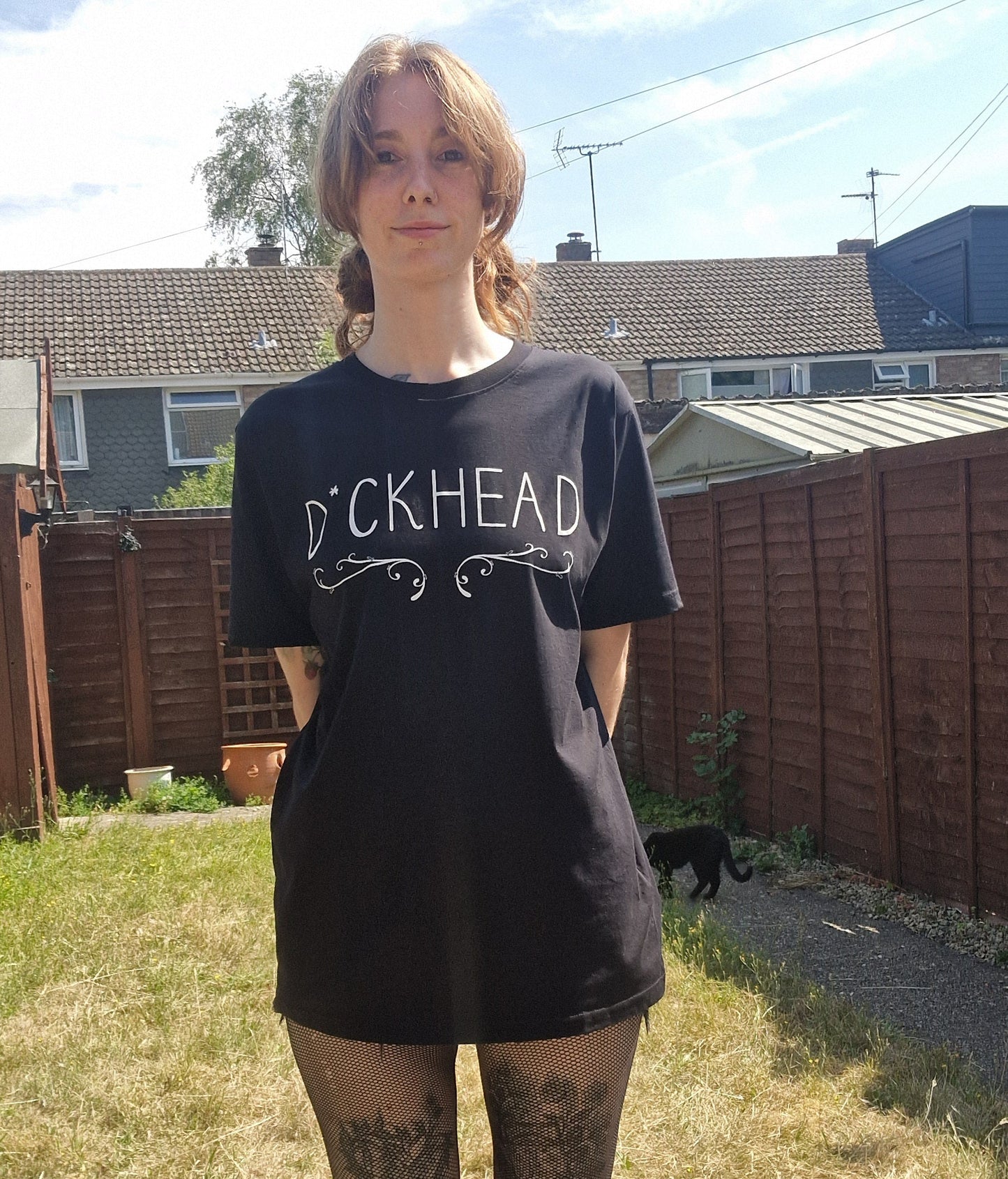 A white woman wears a black t-shirt with the word 'D*ckhead' written across the chest with a filigree inspired design running underneath the writing. Behind her is a green garden and a small black cat.