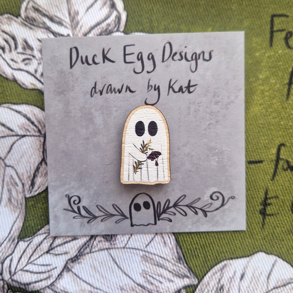 A white ghost pin carrying a drooping red flower and leafy fronds sits on a grey square backing card with black handwriting on that reads 'Duck Egg Designs drawn by Kat'. `Behind the backing card you can see a rich green fabric with a white and black plant design.
