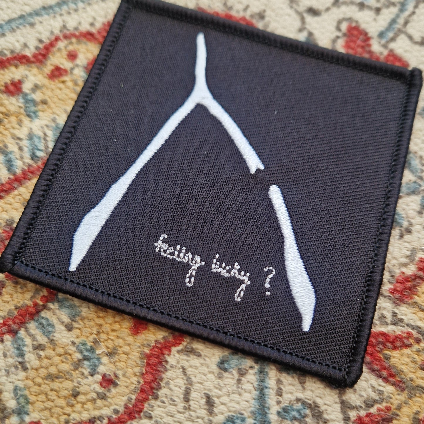 A square black embroidered patch sits on a patterned fabric background. The patch has a white broke wishbone design with the words 'feeling lucky?' underneath it.