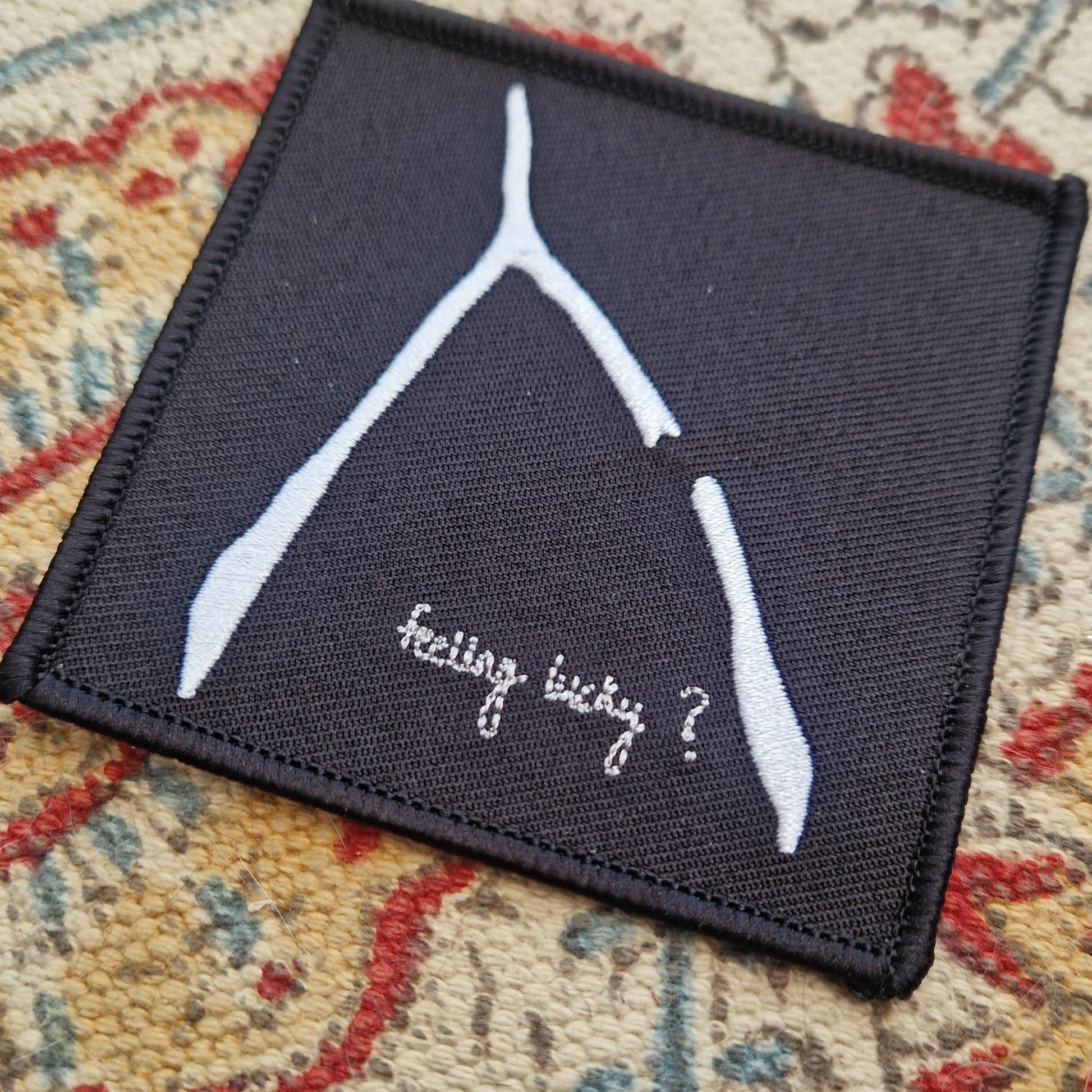 A square black embroidered patch sits on a patterned fabric background. The patch has a white broke wishbone design with the words 'feeling lucky?' underneath it.