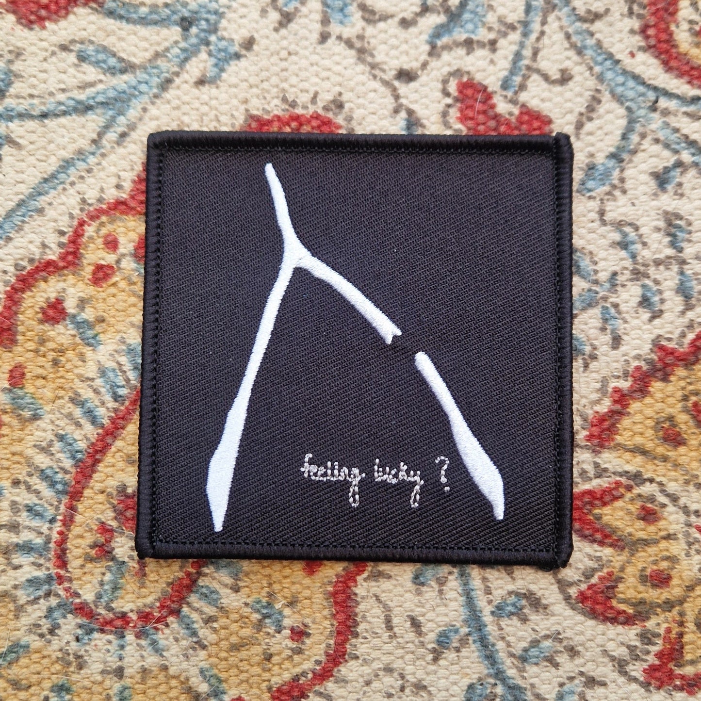 A square black embroidered patch sits on a patterned fabric background. The patch has a white broke wishbone design with the words 'feeling lucky?' underneath it.