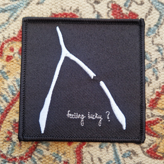 A square black embroidered patch sits on a patterned fabric background. The patch has a white broke wishbone design with the words 'feeling lucky?' underneath it.
