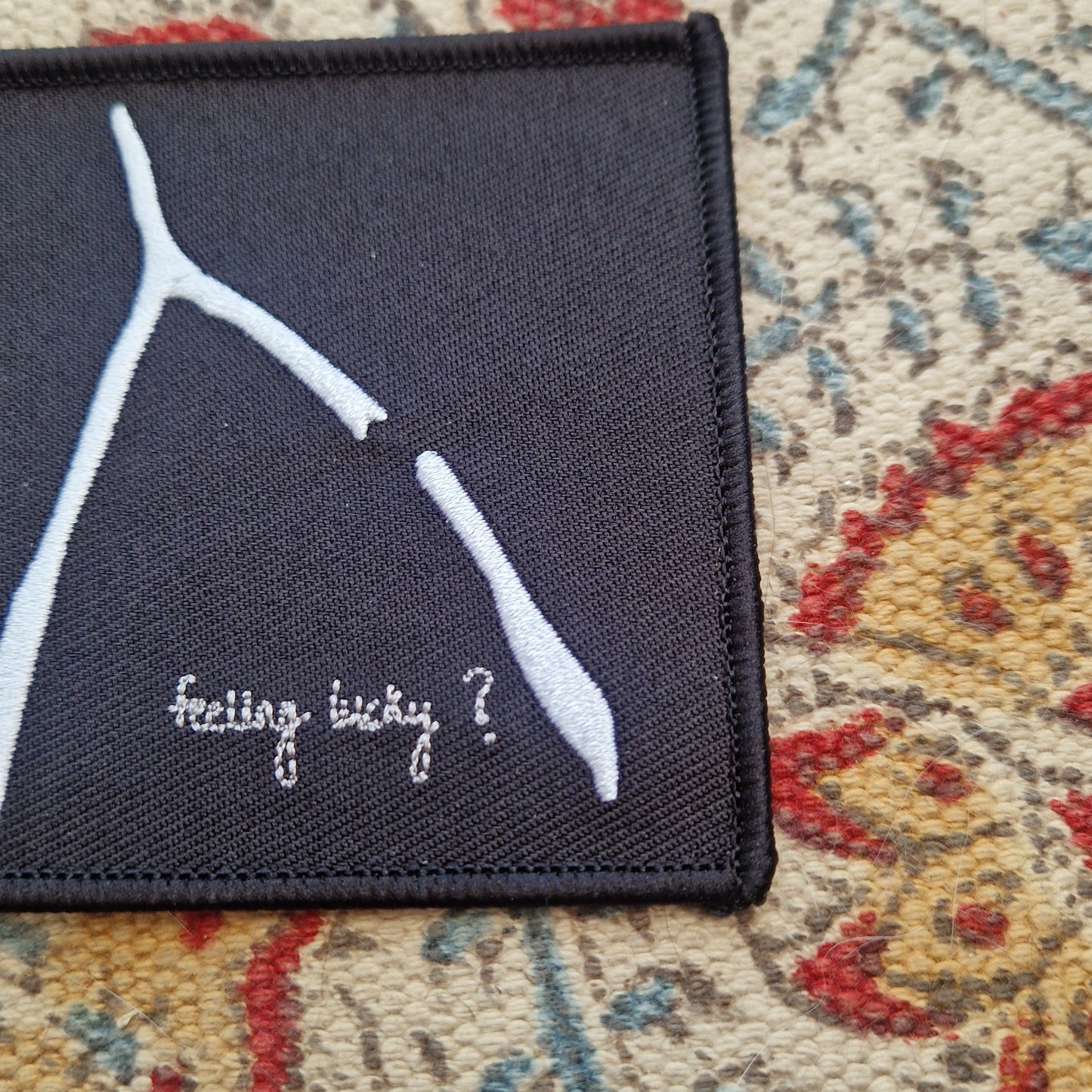 A square black embroidered patch sits on a patterned fabric background. The patch has a white broke wishbone design with the words 'feeling lucky?' underneath it.