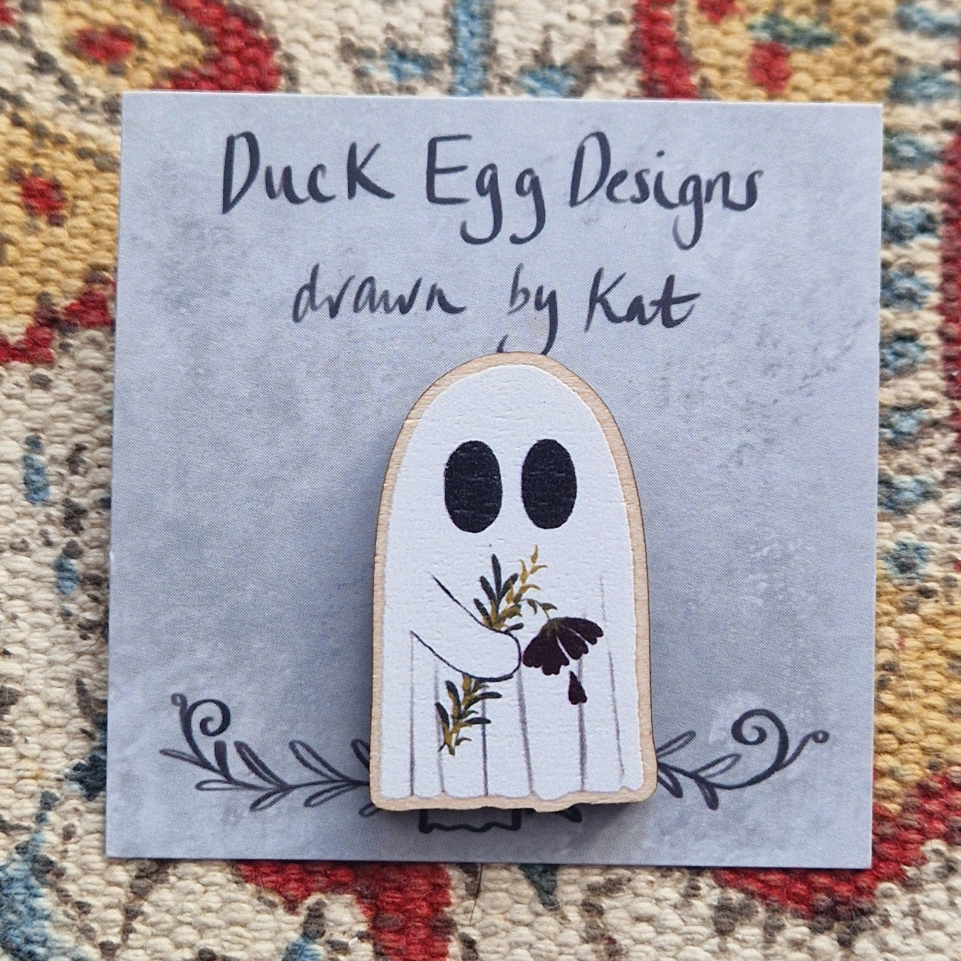 A white ghost carrying a deep red flower and leafy vines pin badge on a grey backing card.