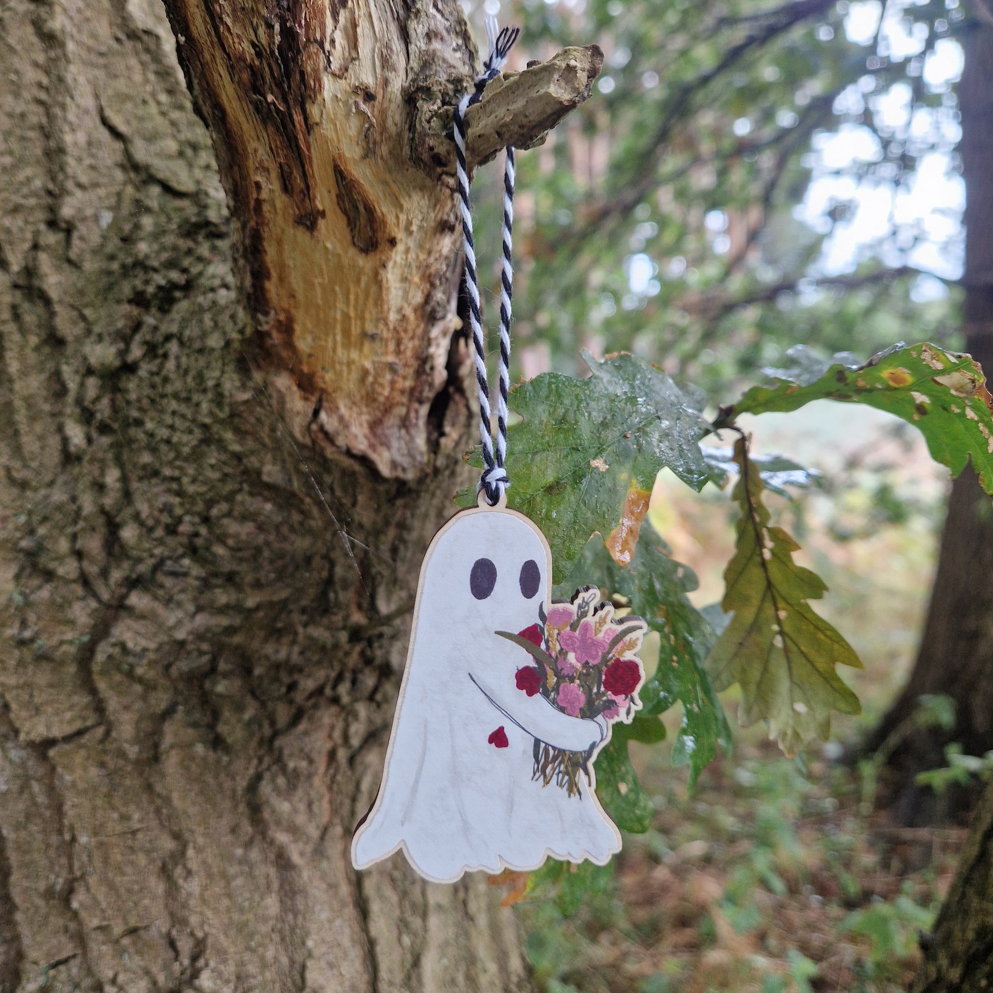 A ghost carrying a bouquet of pink and red flowers printed on wood hangs from a black and white string from a green leaved tree.