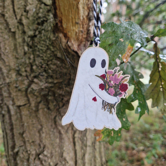 A ghost carrying a bouquet of pink and red flowers printed on wood hangs from a black and white string from a green leaved tree.