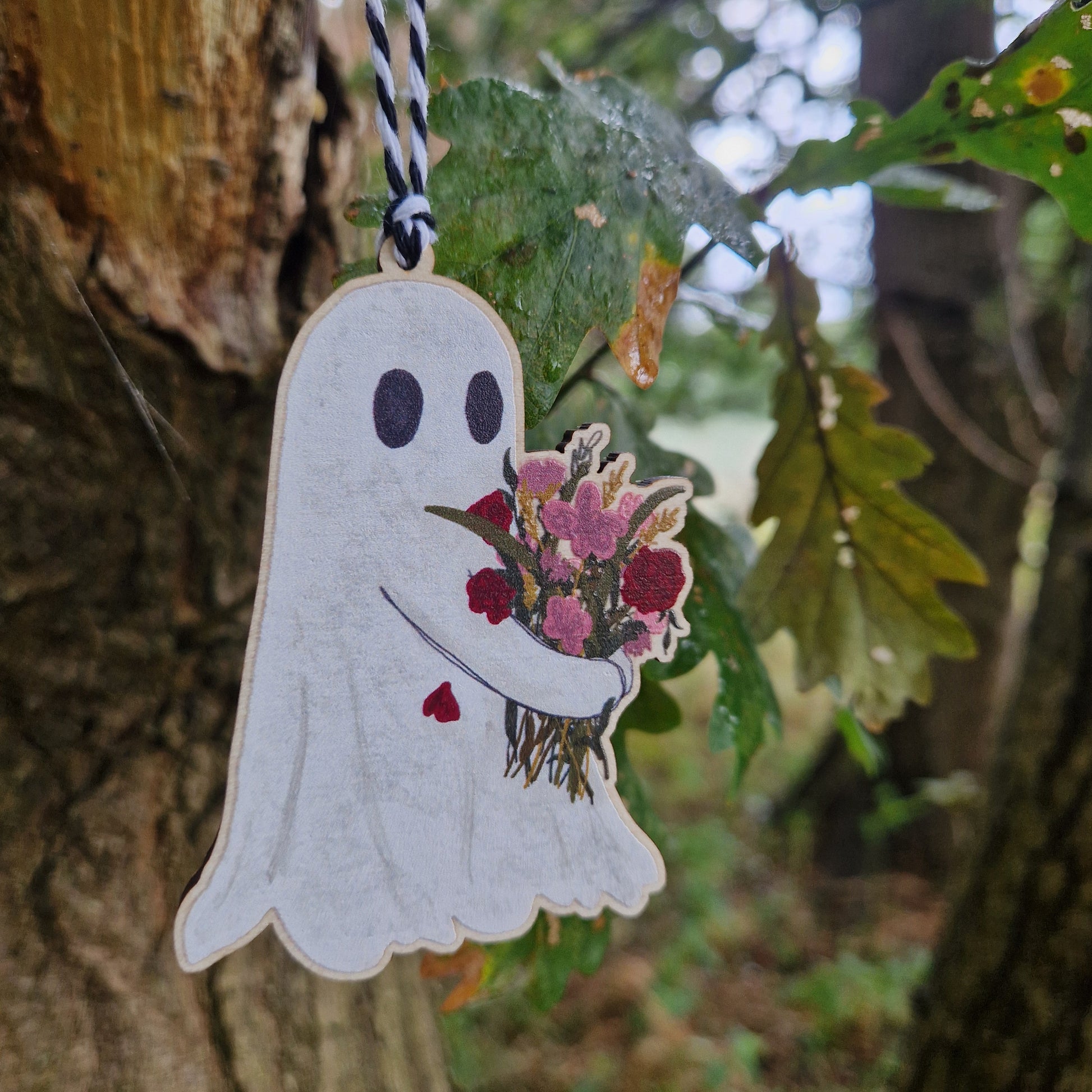 Close up of a ghost carrying a bouquet of pink and red flowers printed on wood hangs from a black and white string from a green leaved tree.