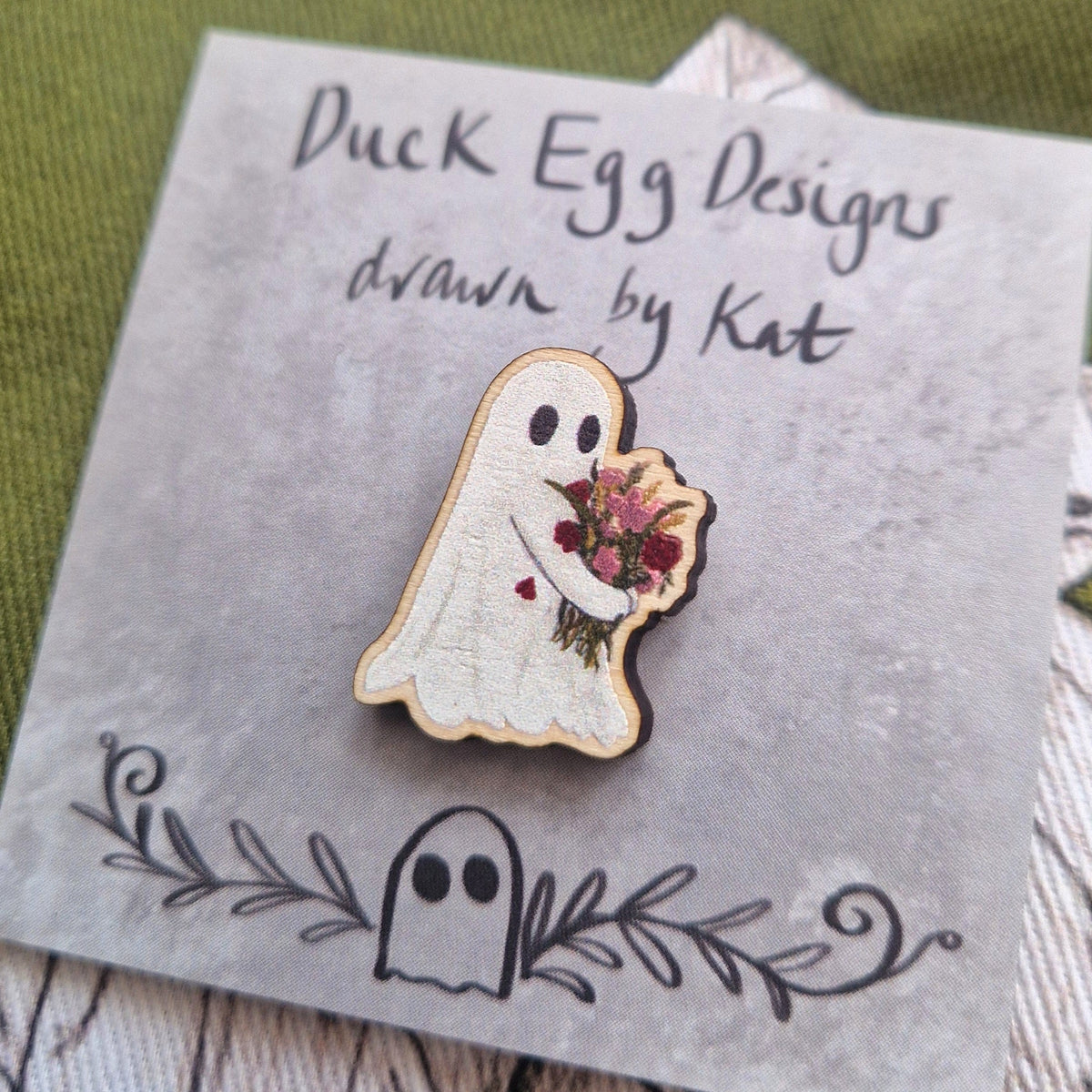 Flower carrying ghost pin badge sits on a grey backing card with the words 'Duck Egg Designs drawn by Kat' in black handwriting. Behind the backing card is a rich green fabric with a white and black plant illustration design.