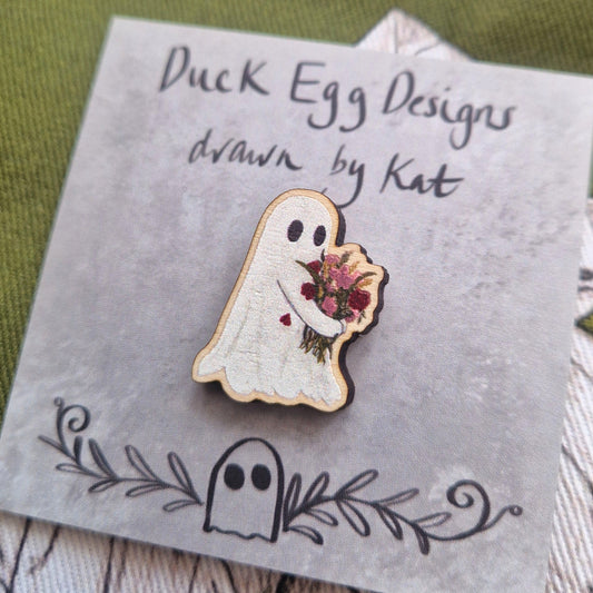 Flower carrying ghost pin badge sits on a grey backing card with the words 'Duck Egg Designs drawn by Kat' in black handwriting. Behind the backing card is a rich green fabric with a white and black plant illustration design.