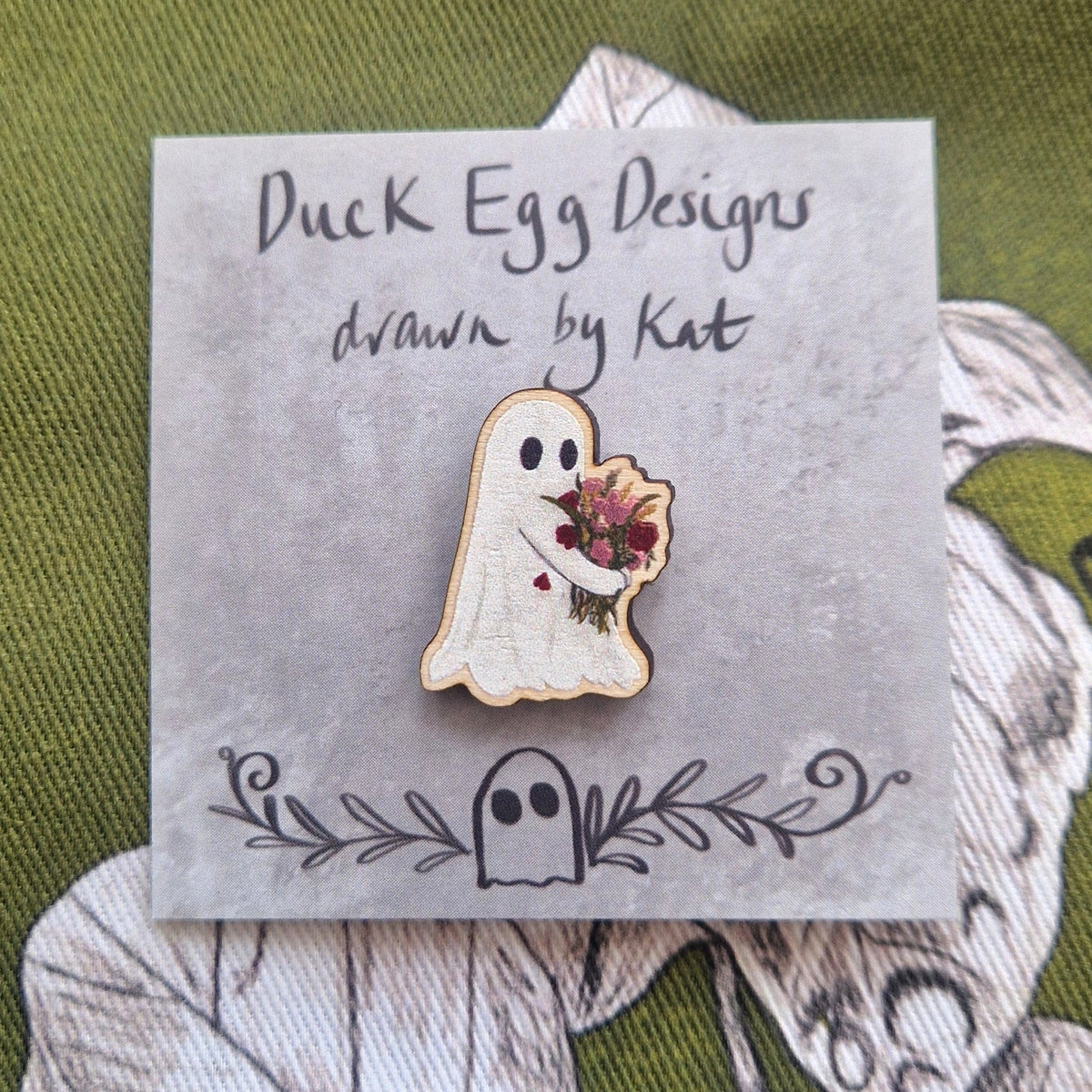 A small pin of a ghost carrying a bouquet of pink and red flowers sits on a grey backing card with the words 'Duck Egg Designs, drawn by Kat' in black handwriting. Behind the grey backing card is a rich green fabric with a white and black leafy design.