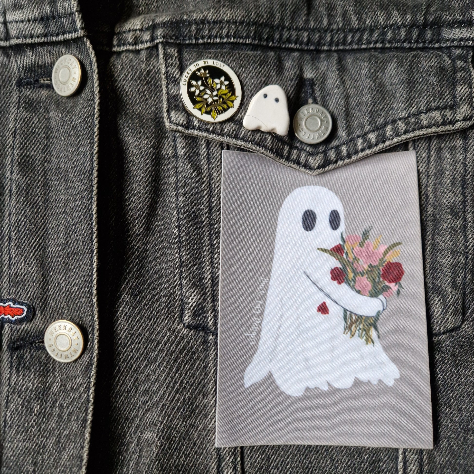A grey denim jacket with ghost pin badge and flower pin on a pocket flap above a grey rectangular patch featuring a white ghost carrying a bouquet of pink and red flowers.