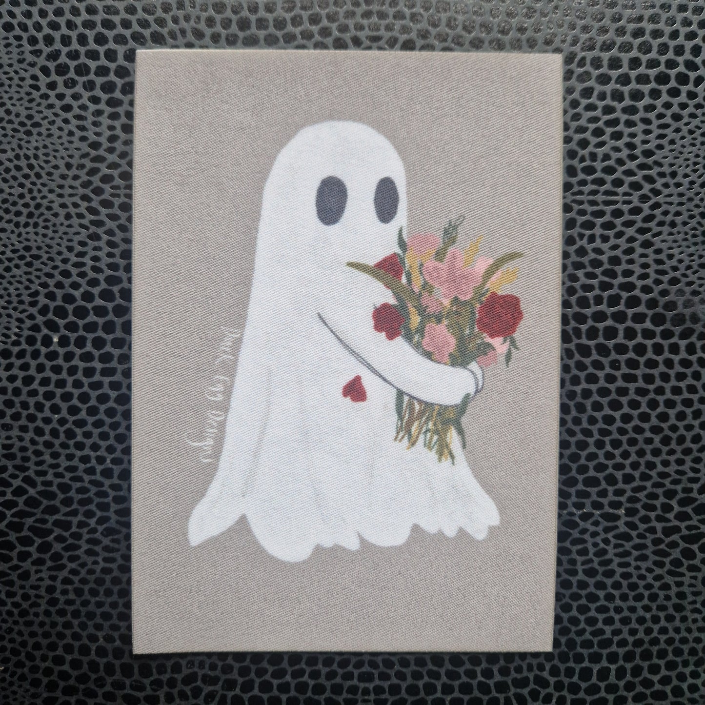 A grey rectangular patch featuring a white ghost carrying a bouquet of flowers. Behind the patch is a black and grey  printed snakeskin bookcover.