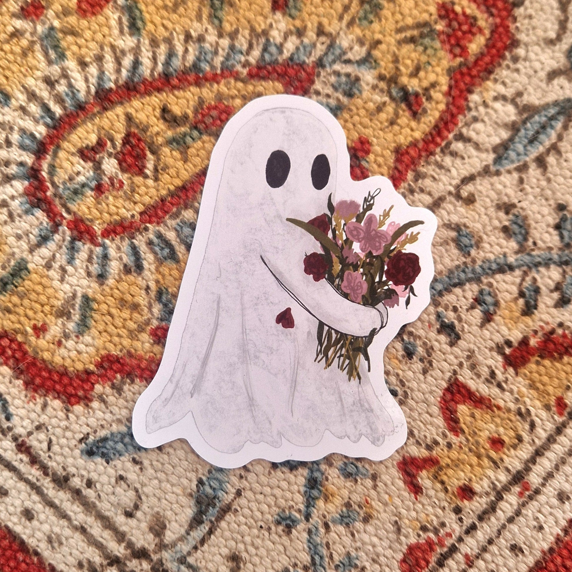 A sticker featuring a white ghost carrying a bouquet of pink and red flowers sits on a patterned fabric.