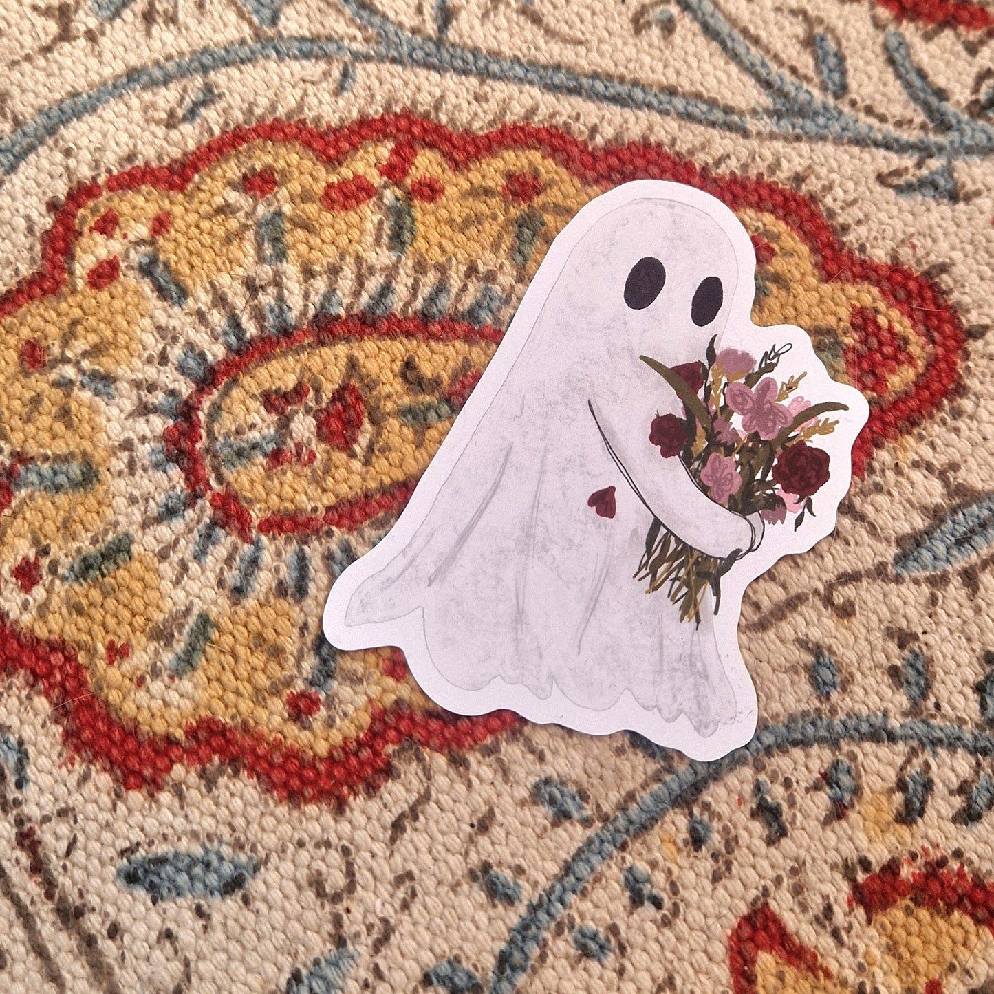 A sticker featuring a white ghost carrying a bouquet of pink and red flowers sits on a patterned fabric.