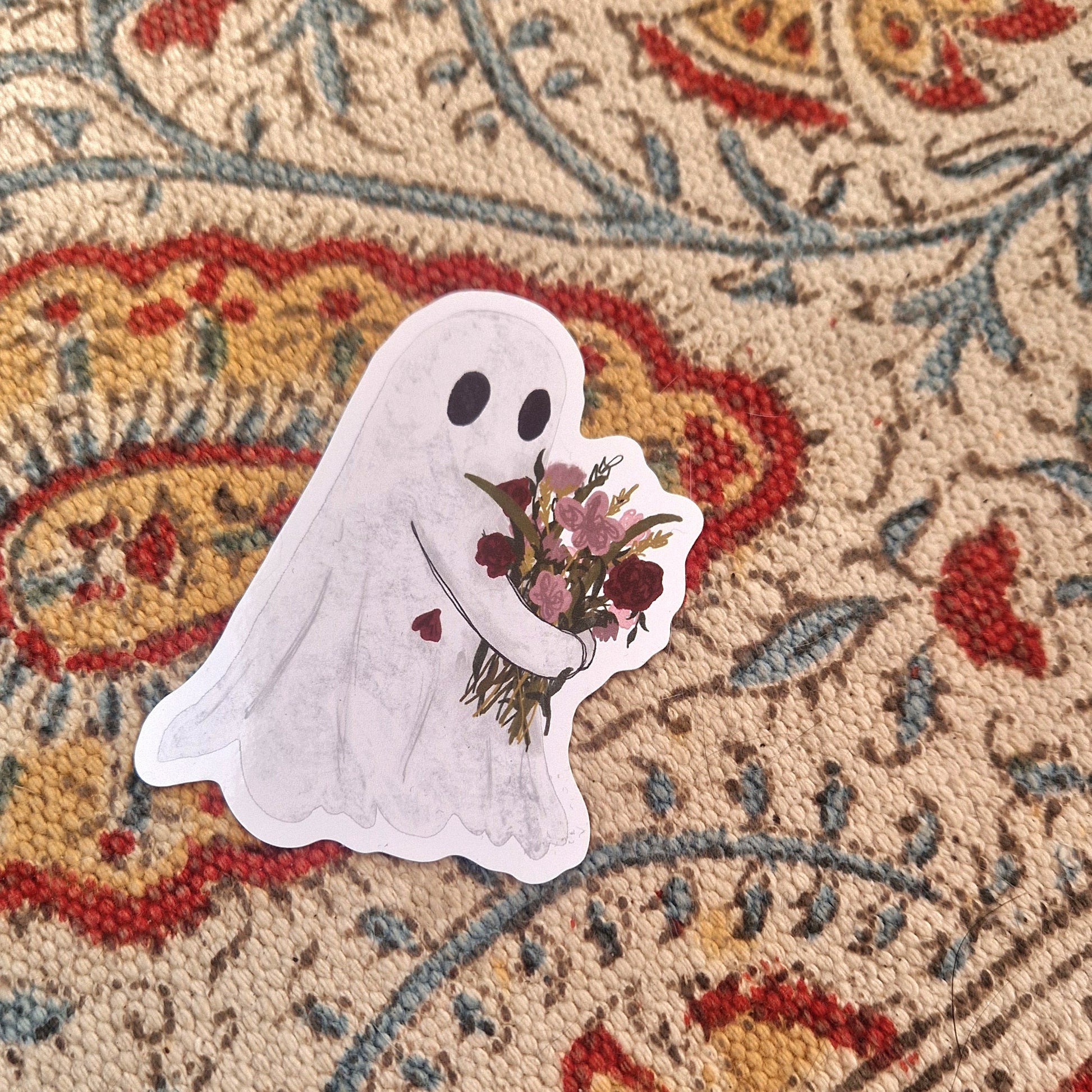 A sticker featuring a white ghost carrying a bouquet of pink and red flowers sits on a patterned fabric.