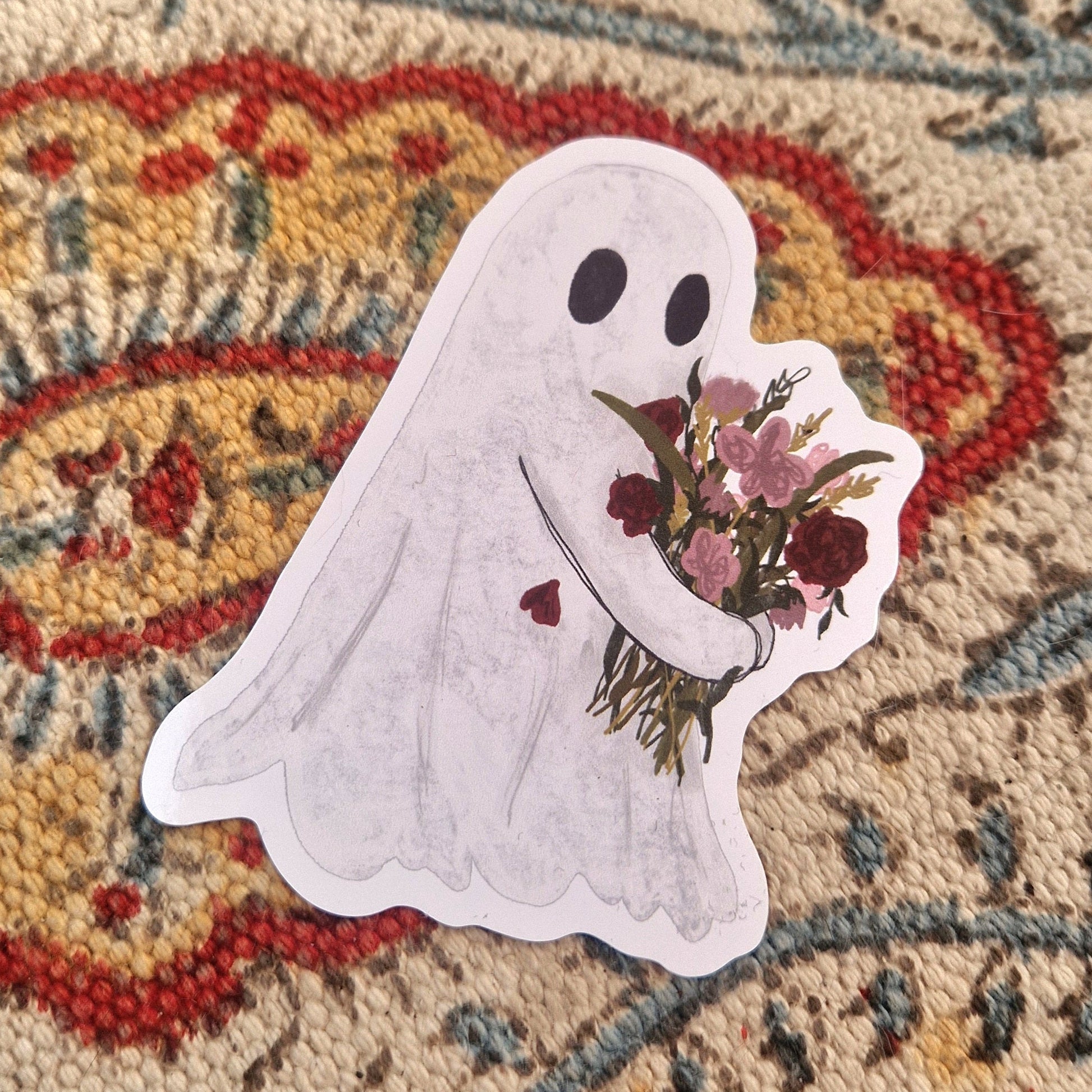 A sticker featuring a white ghost carrying a bouquet of pink and red flowers sits on a patterned fabric.