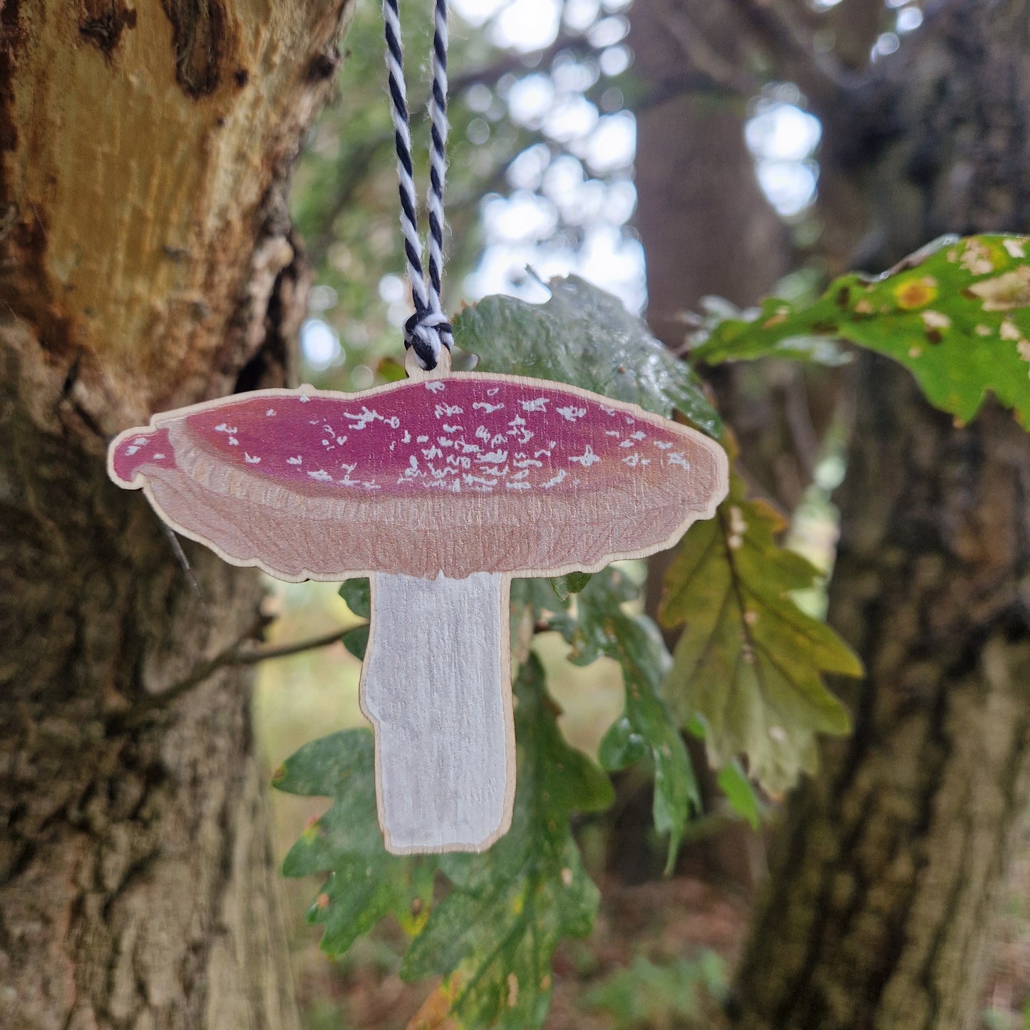 A red and white toadstool printed on woods hangs from a green leaved tree from a white and black string.
