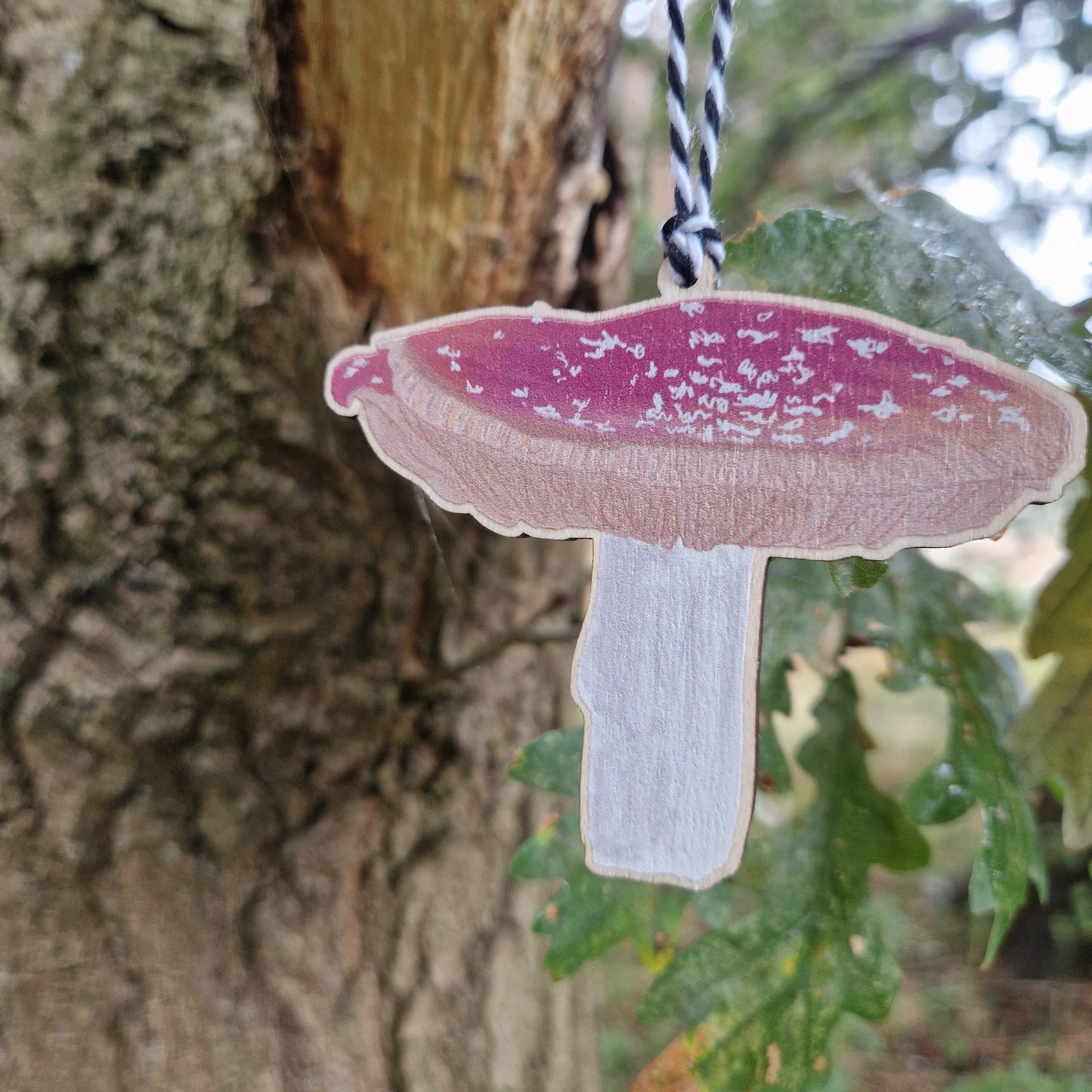 A close up view of a red and white toadstool printed on woods hangs from a green leaved tree from a white and black string.