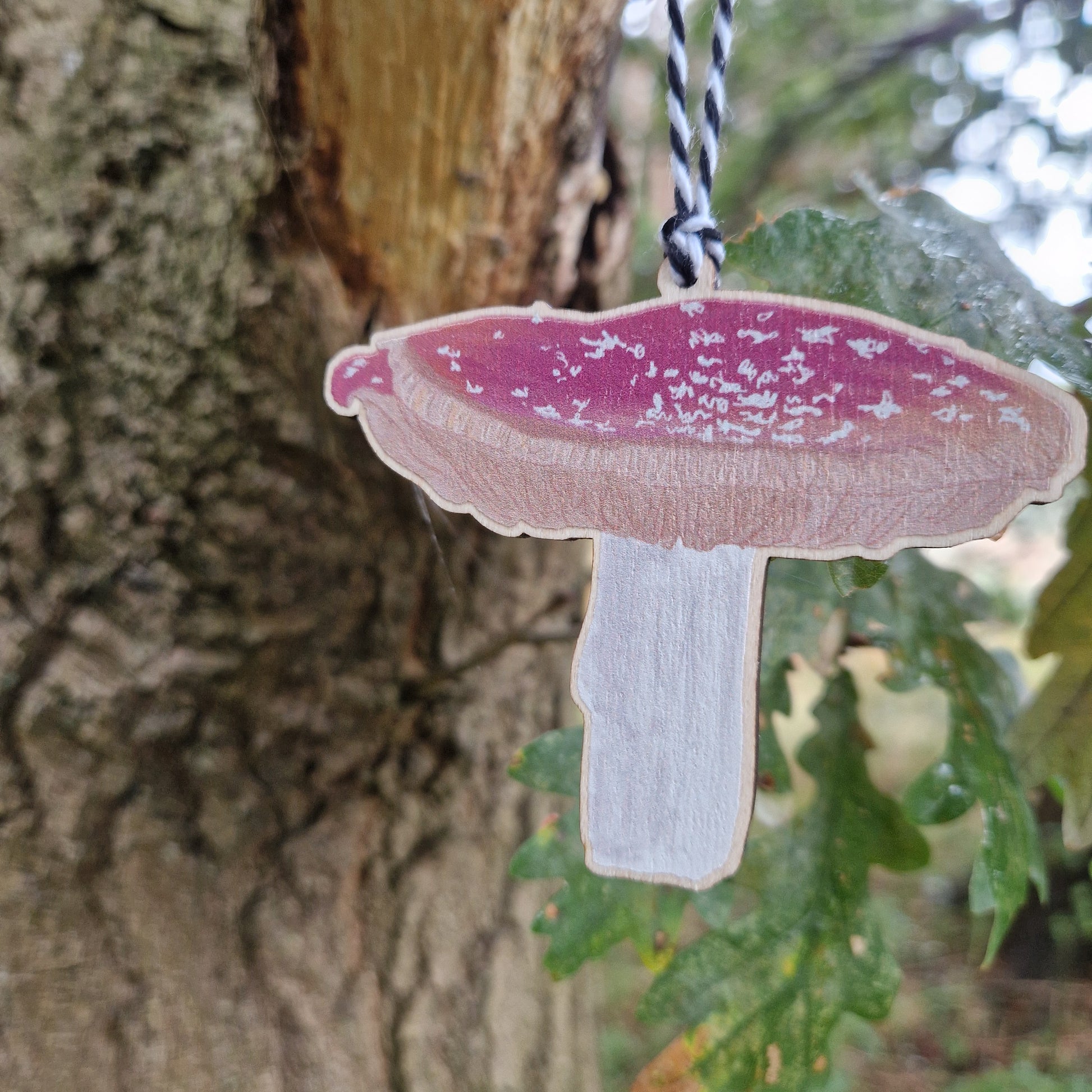 A close up view of a red and white toadstool printed on woods hangs from a green leaved tree from a white and black string.