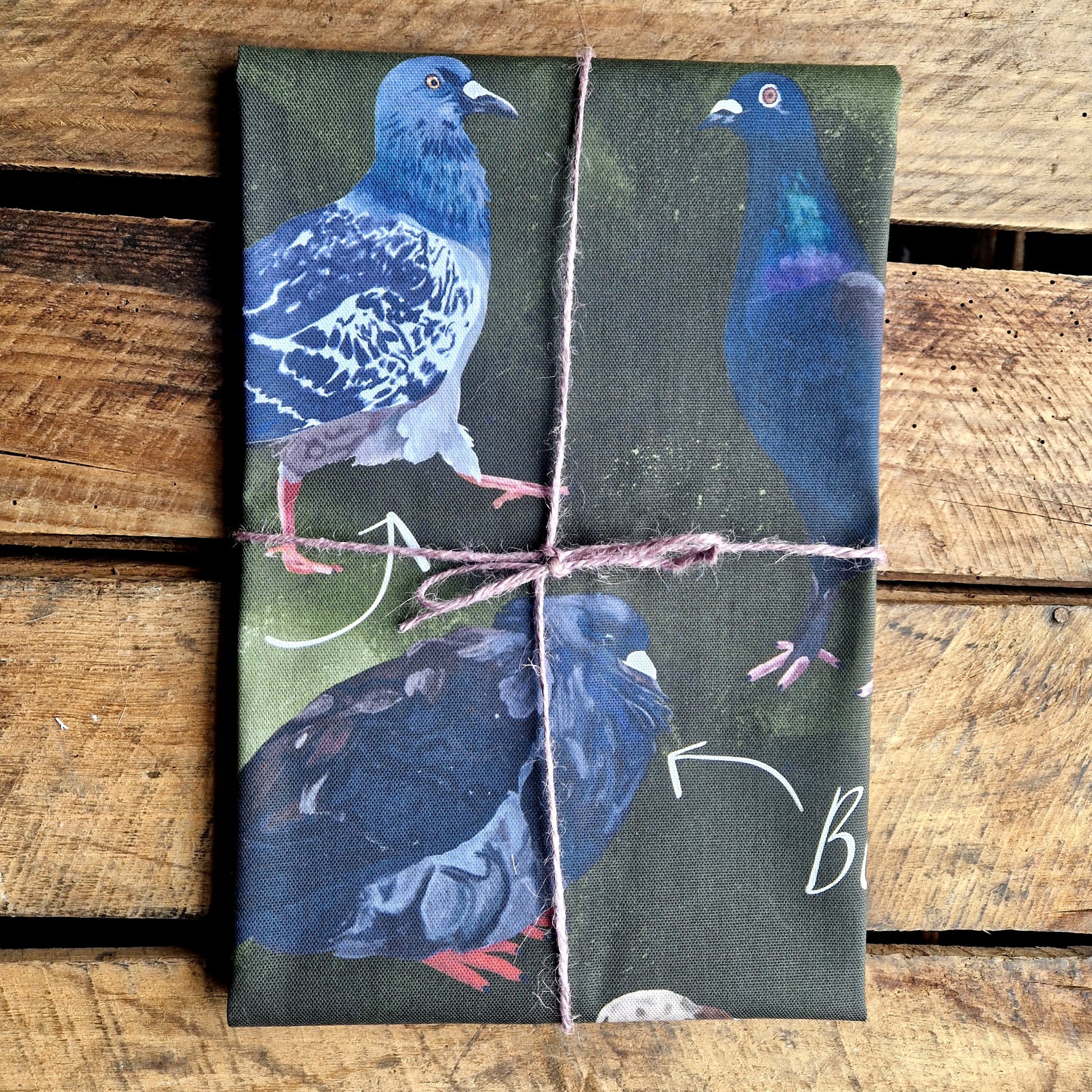 A folded green tea towel with three pigeons visible sits on a wooden plank background. The tea towel is wrapped in a lilac macrame twine.