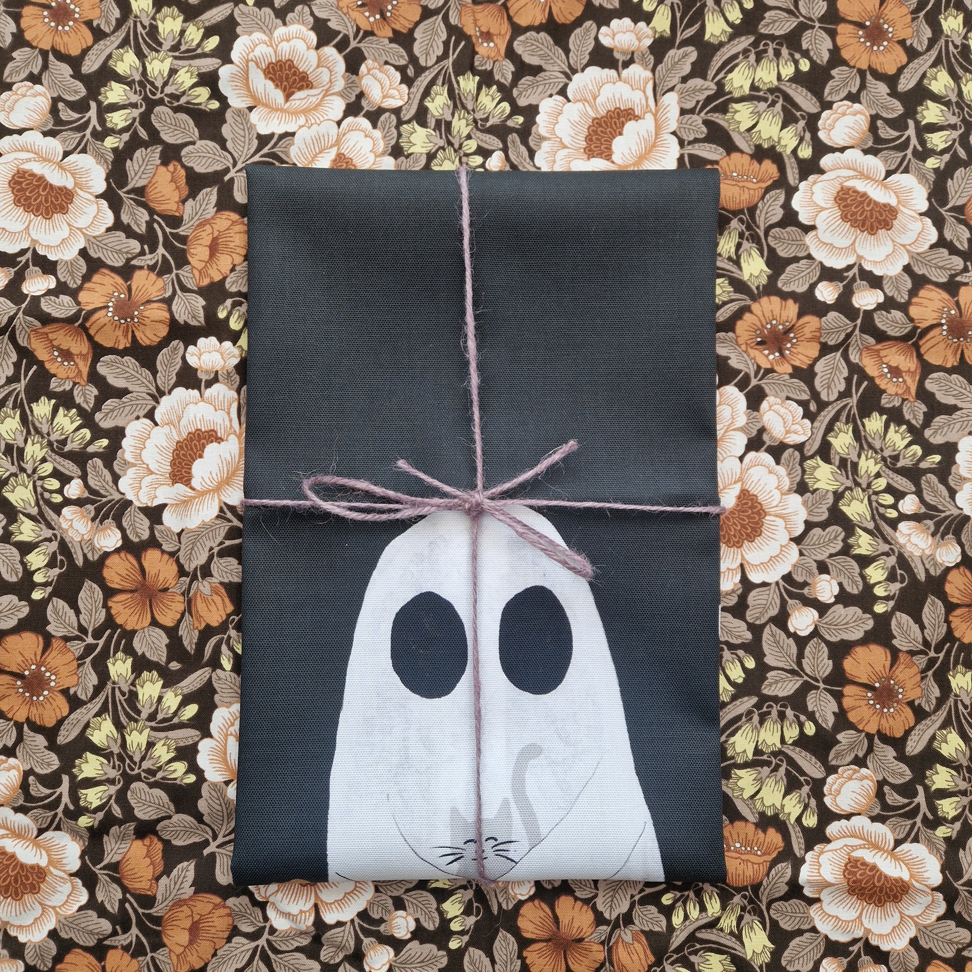 A folded black tea towel lies on a floral patterned fabric wrapped in a purple string. You can see the top of a ghost holding a grey cat on the bottom half of the tea towel.