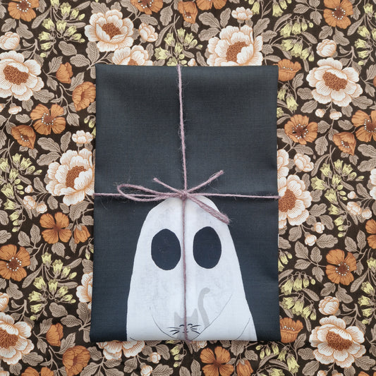 A folded black tea towel lies on a floral patterned fabric wrapped in a purple string. You can see the top of a ghost holding a grey cat on the bottom half of the tea towel.