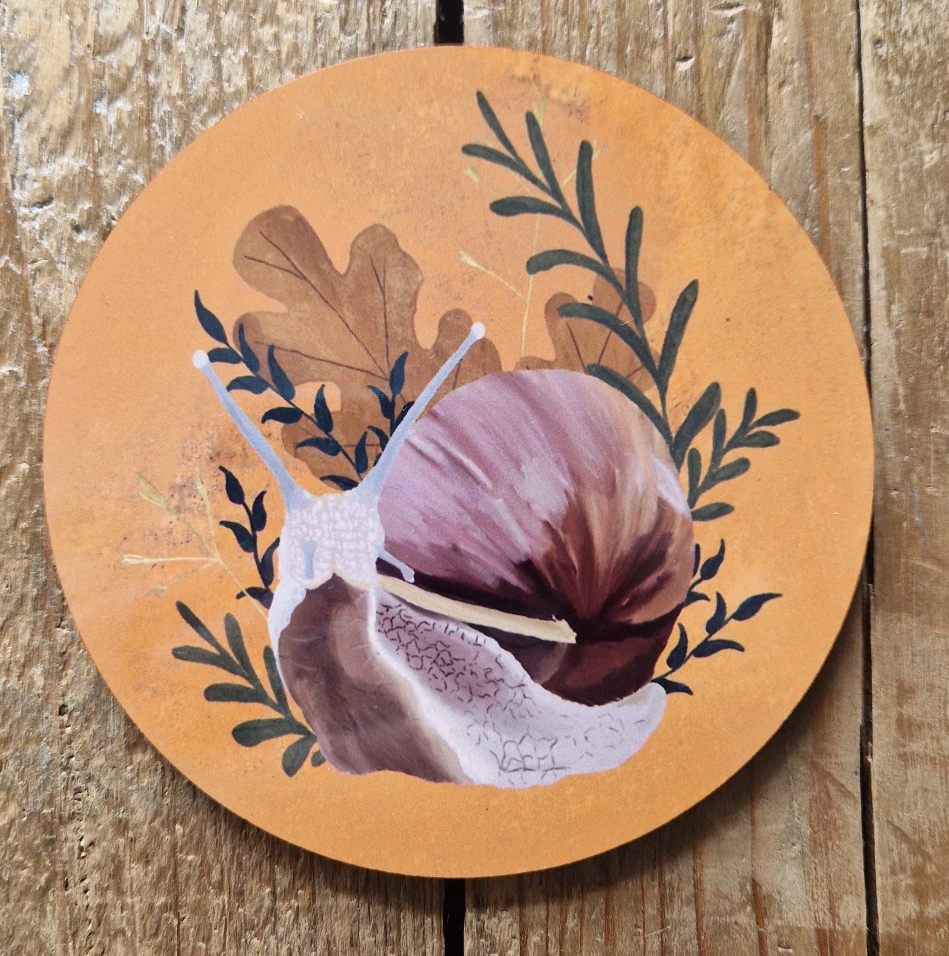 A round snail coaster featuring a pink, yellow and brown snail surrounded by green leafy vines, brown oak leaves and yellow dried grasses. Behind the coaster is a wooden plank background.