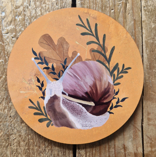 A round snail coaster featuring a pink, yellow and brown snail surrounded by green leafy vines, brown oak leaves and yellow dried grasses. Behind the coaster is a wooden plank background.