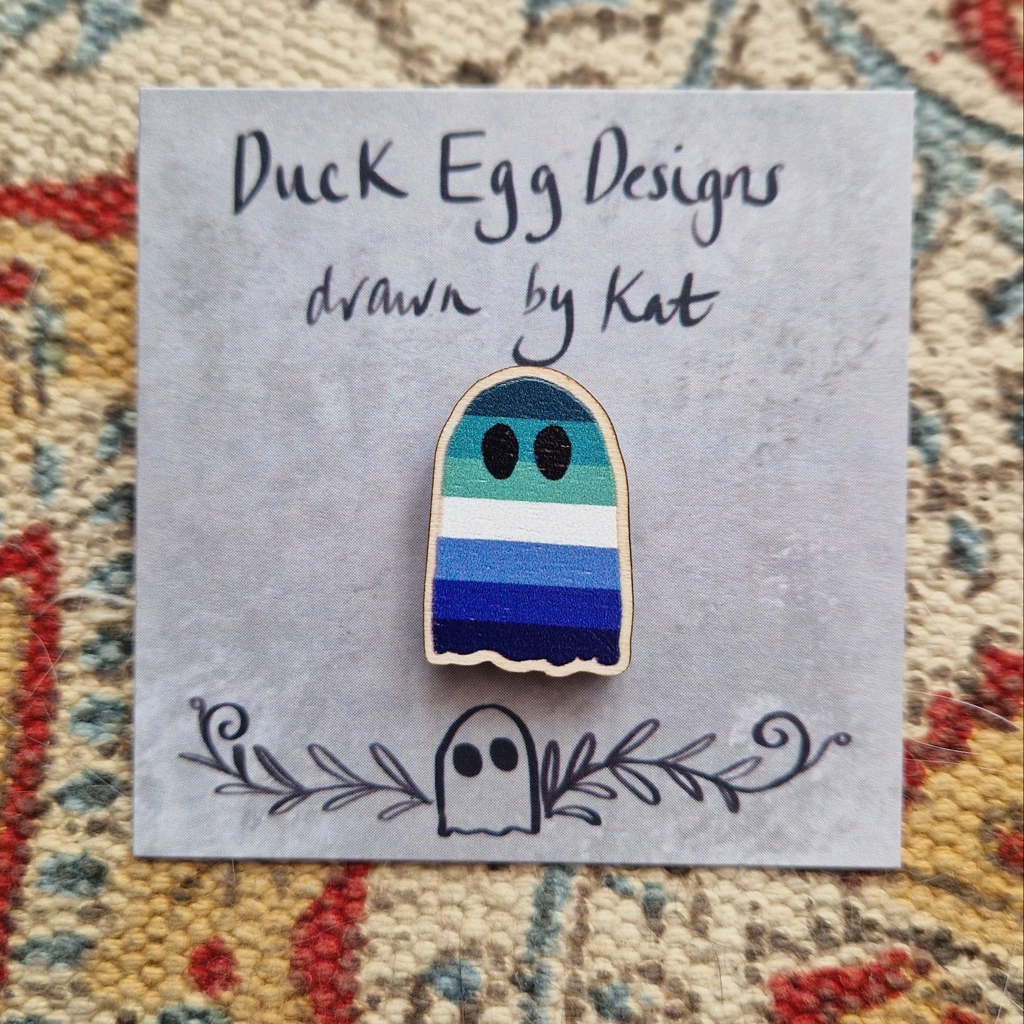 A blue stripey gay pride flag ghost pin sits on a grey backing card with the words 'Duck Egg Designs drawn by Kat' written across the top. Behind the backing card is a patterned fabric background.