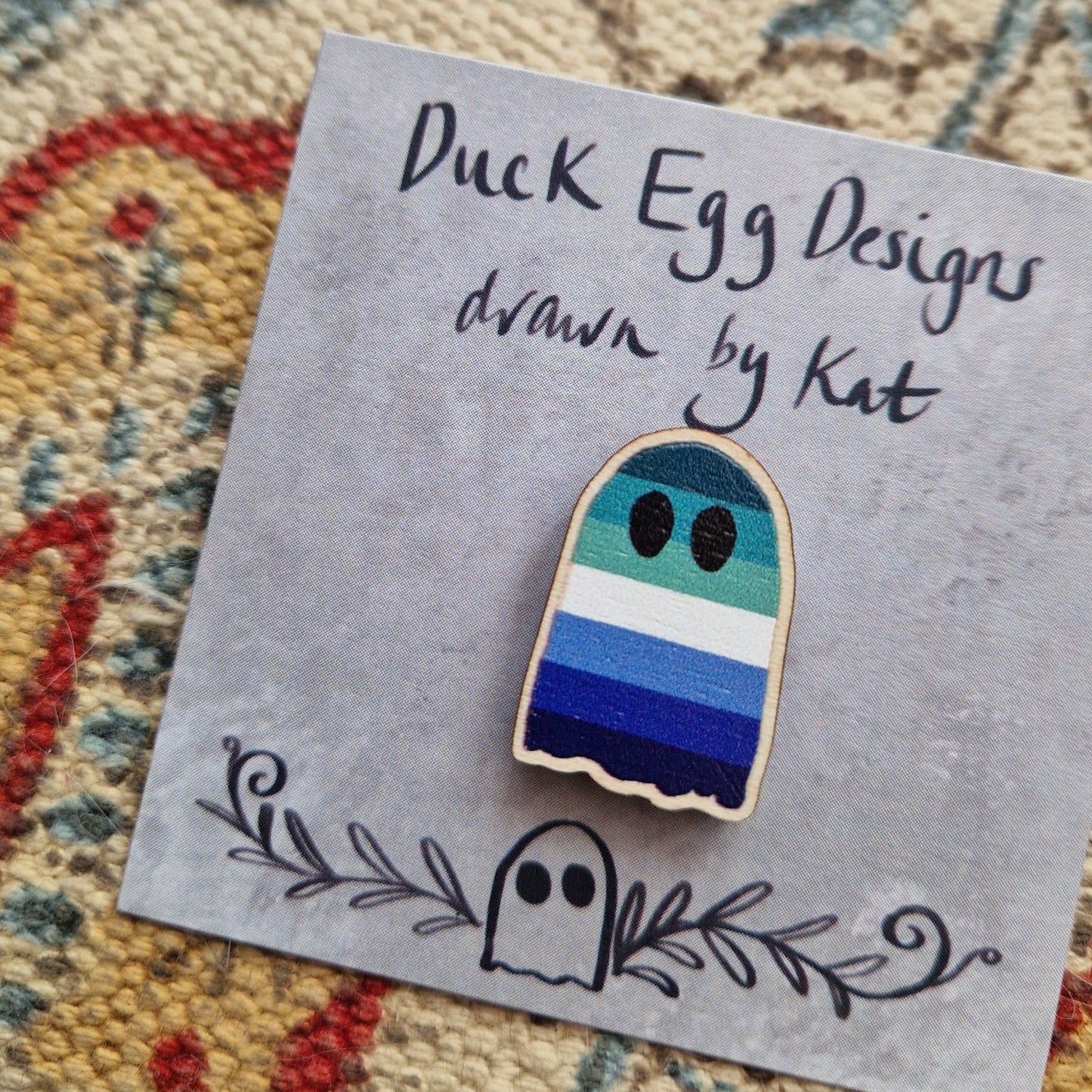 A closer look at the blue, green and white gay p[ride flag ghost pin badge on it's grey backing card showing the texture of the wooden pin.