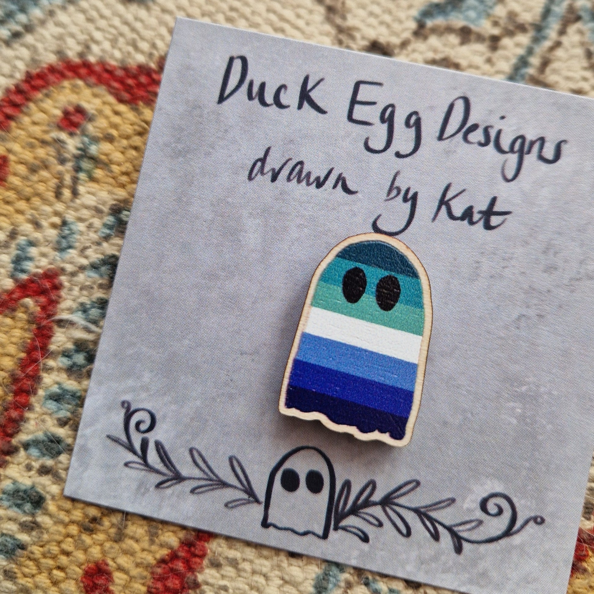 A closer look at the blue, green and white gay p[ride flag ghost pin badge on it's grey backing card showing the texture of the wooden pin.
