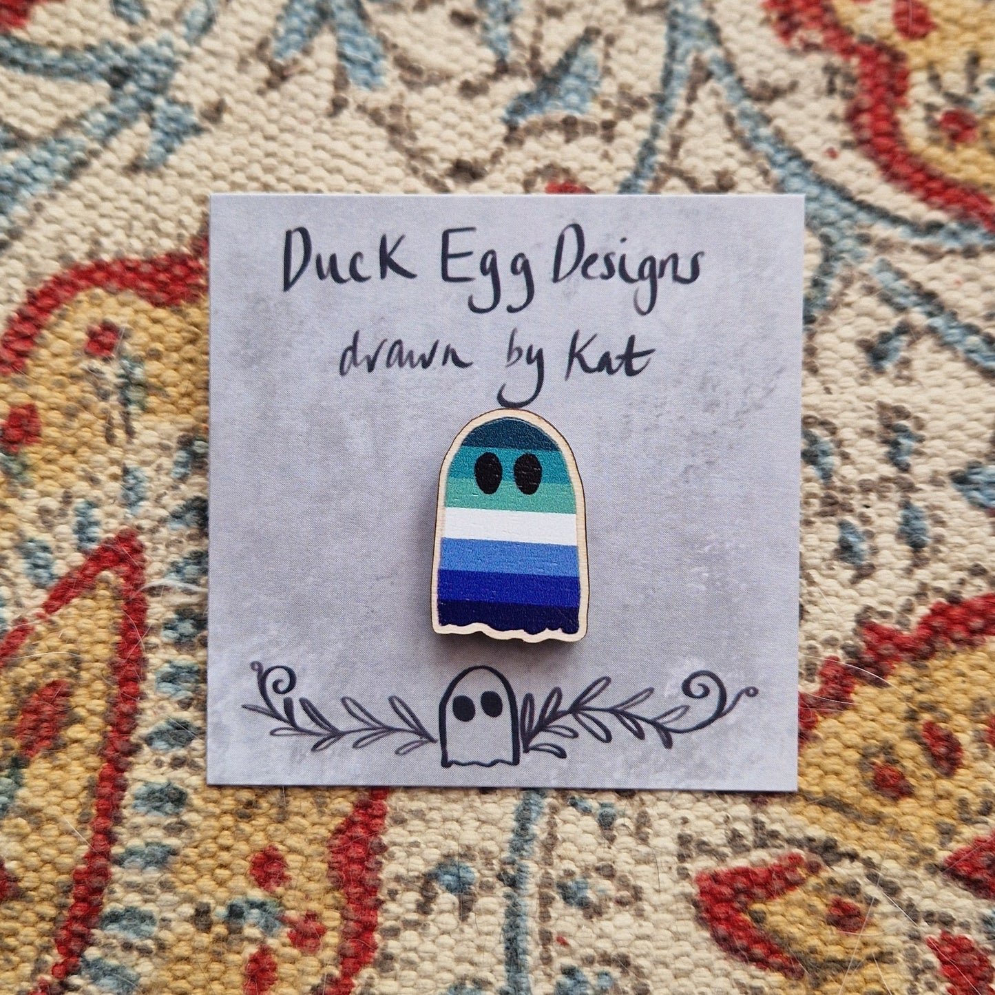 A blue stripey gay pride flag ghost pin sits on a grey backing card with the words 'Duck Egg Designs drawn by Kat' written across the top. Behind the backing card is a patterned fabric background.