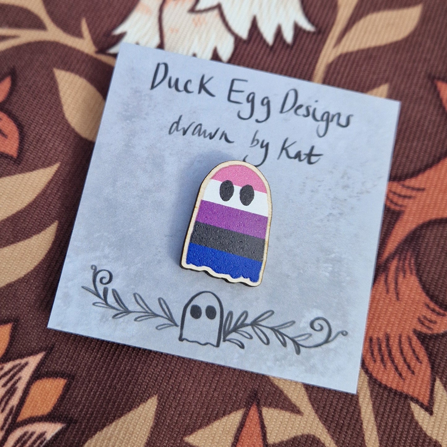 A ghost pin featuring the colourful stripes of the genderfluid flag sits on a grey square backing card with the words 'Duck Egg Designs drawn by Kat' in black across the top. The backing card sits on a brown patterned fabric.