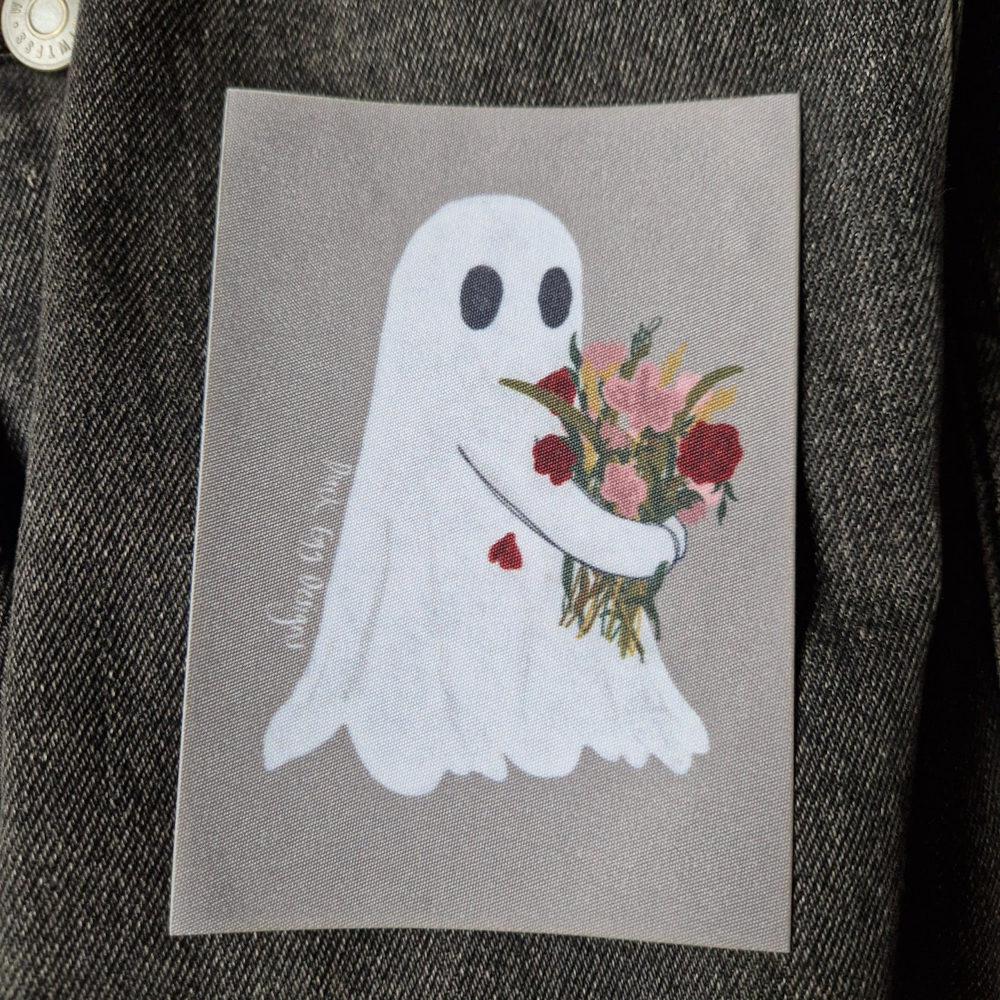 Closer view of the printed  grey rectangular patch featuring a ghost carrying a bouquet of red and pink flowers.