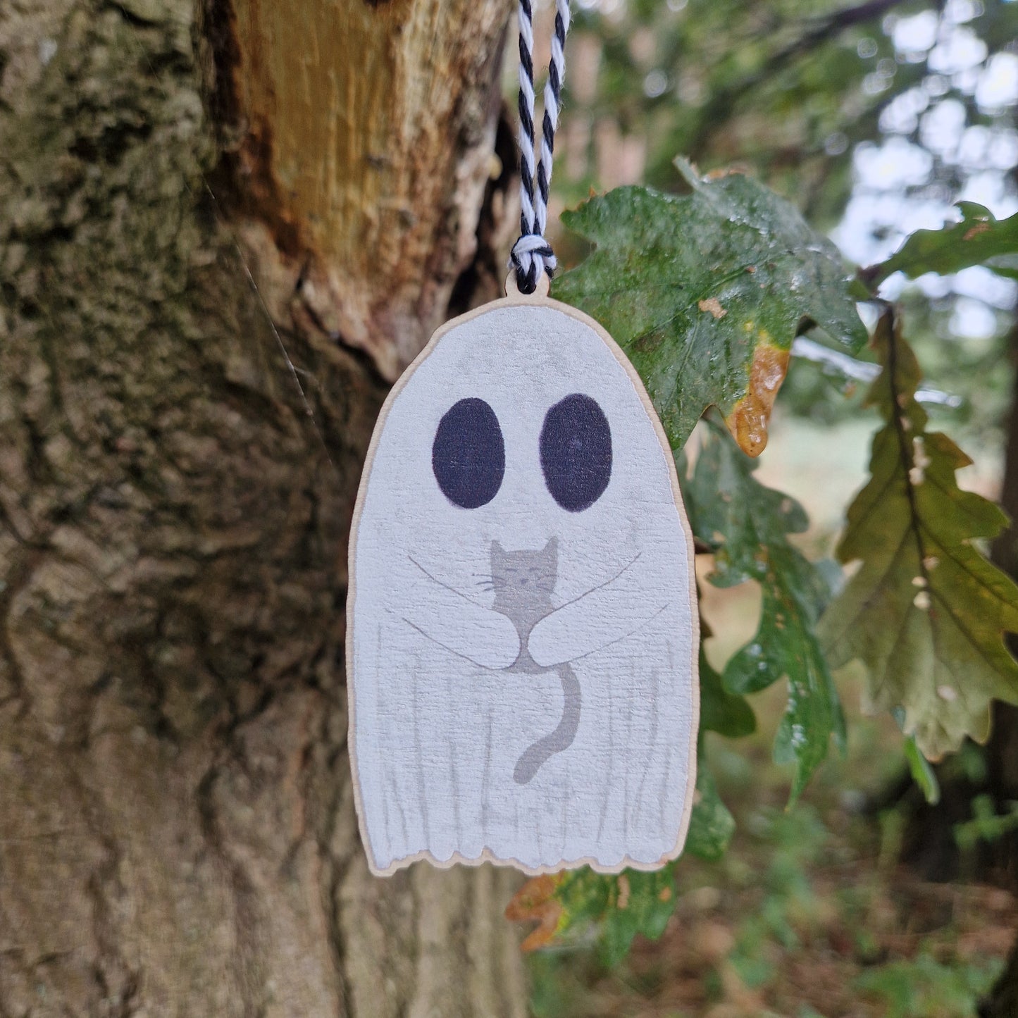 A close up look at the white ghost holding a grey cat hangs by a white and black string from a green leafed tree.