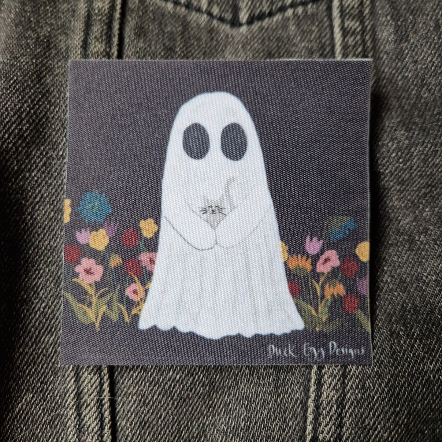 Square black patch featuring a ghost carrying a grey cat in a field of yellow, blue, red and pink flowers. Behind the patch you can see a grey denim jacket.