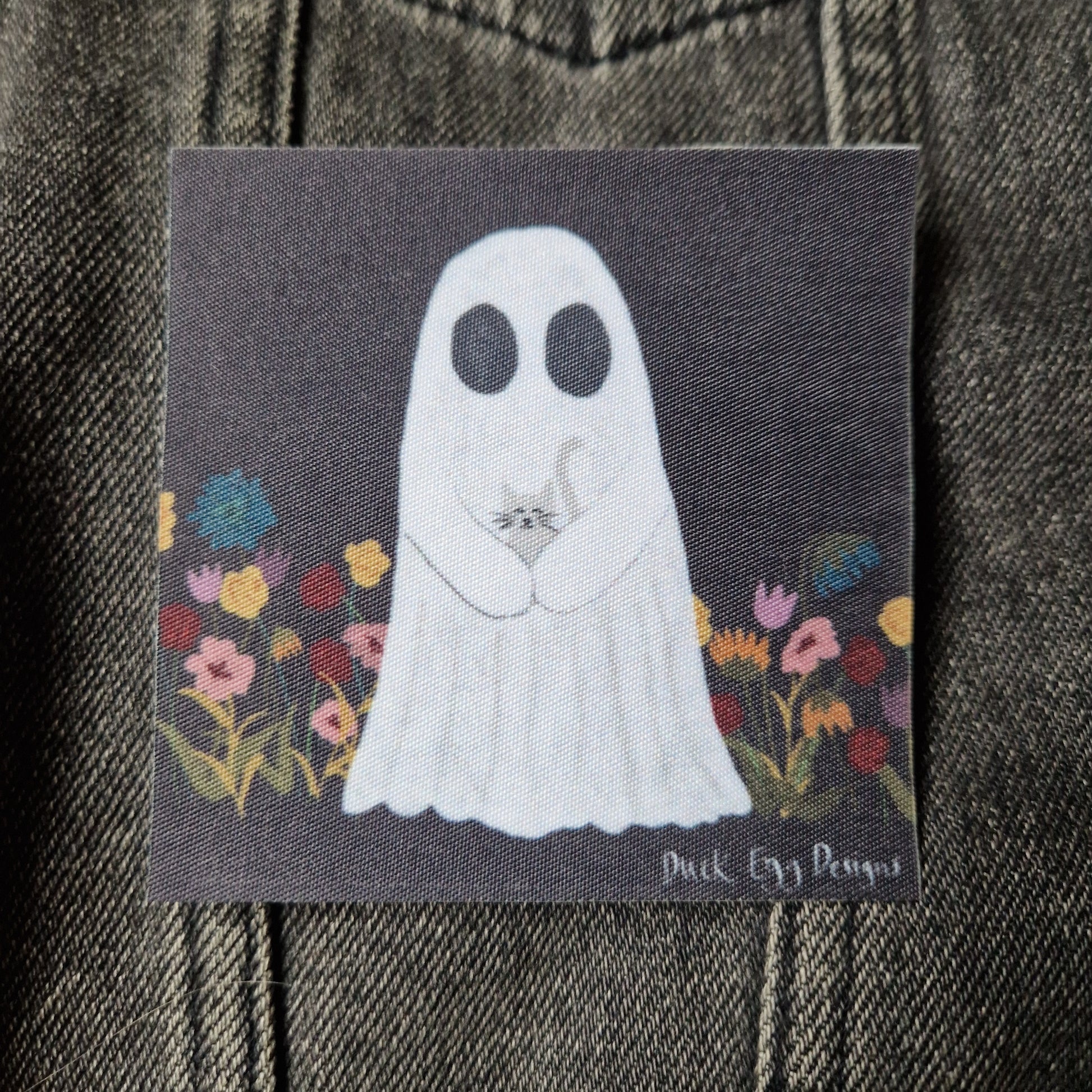 Square black patch featuring a ghost carrying a grey cat in a field of yellow, blue, red and pink flowers. Behind the patch you can see a grey denim jacket.