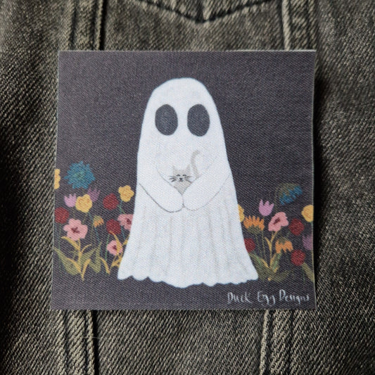 Square black patch featuring a ghost carrying a grey cat in a field of yellow, blue, red and pink flowers. Behind the patch you can see a grey denim jacket.