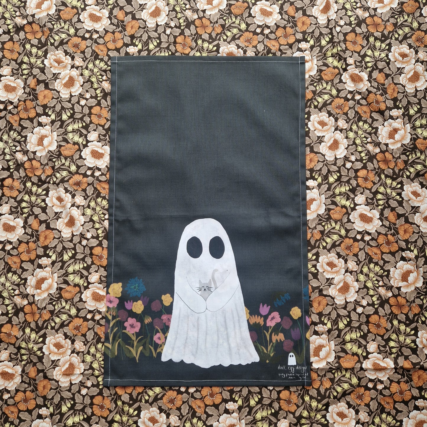 A black tea towel with a ghost holding a grey cat on the bottom half, surrounded by colourful flowers. 