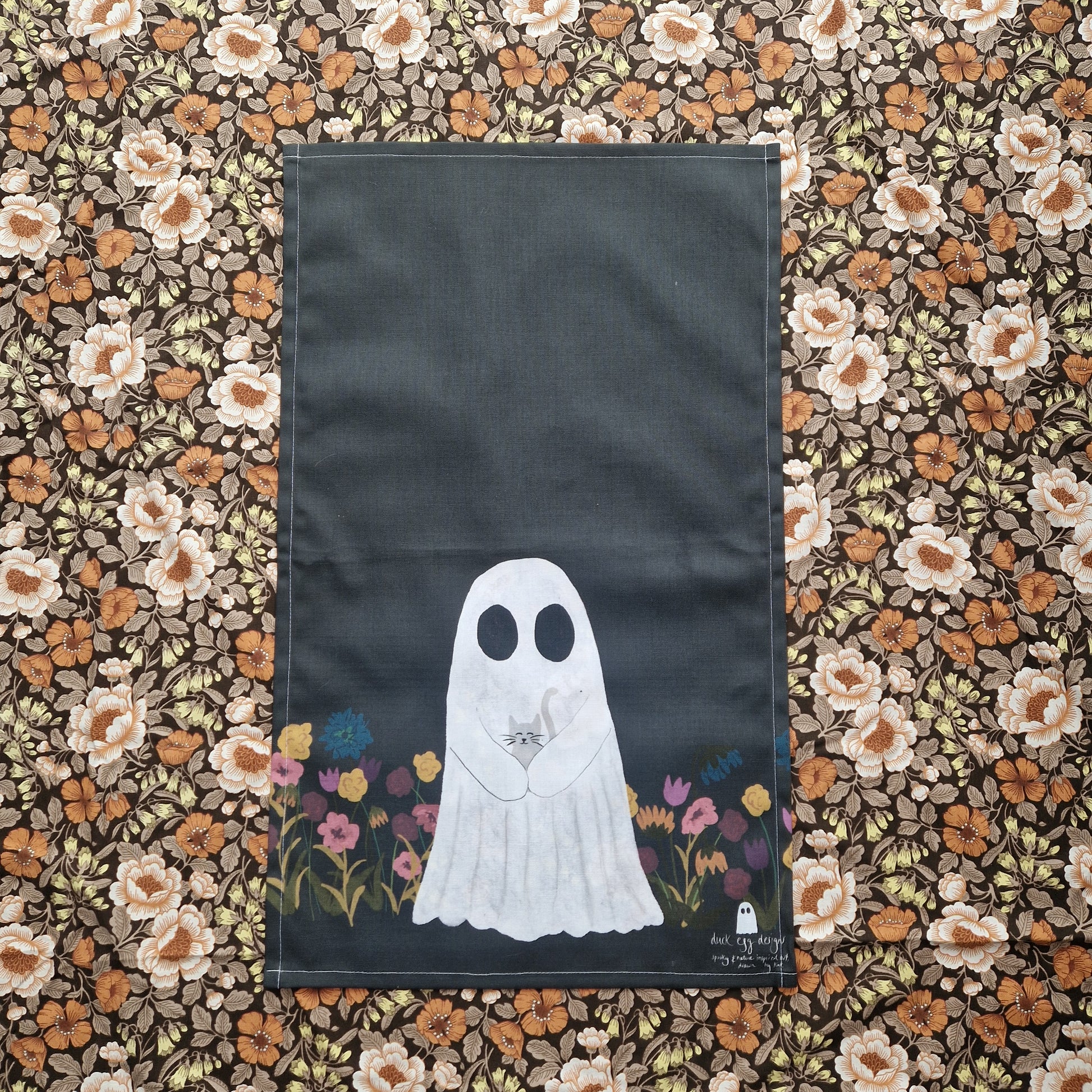 A black tea towel with a ghost holding a grey cat on the bottom half, surrounded by colourful flowers. 