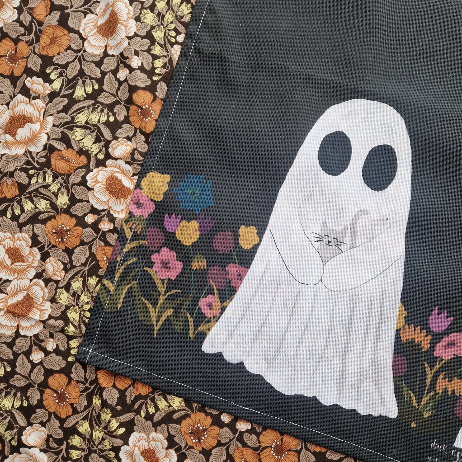 A closer look at the tea towel showing the colourful flowers and the ghost holding a grey cat in the bottom half of the design. Behind the tea towel is a floral patterned fabric.