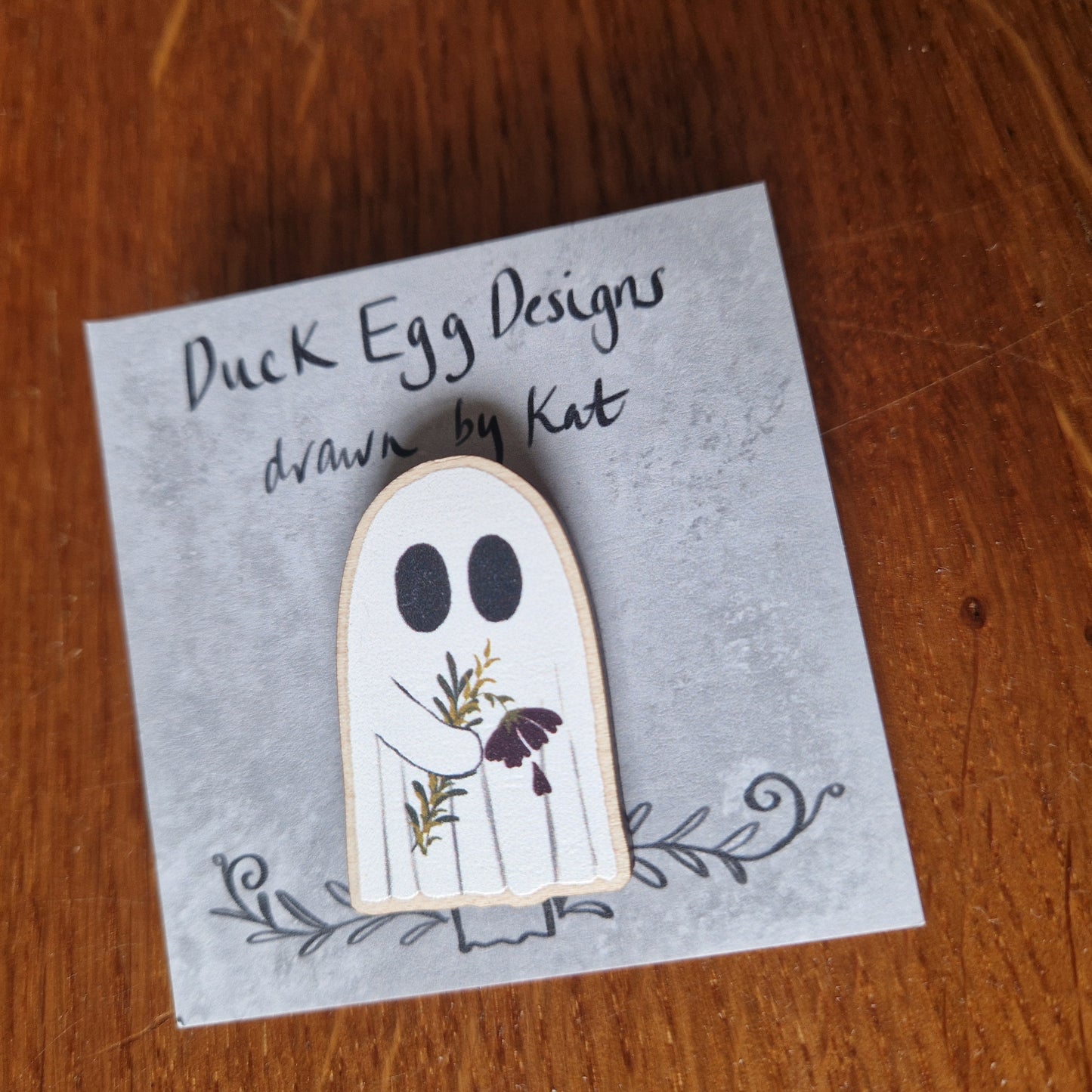 A white ghost pin badge sits on a grey backing card with the words 'Duck Egg Designs drawn by Kat' across the top. The ghost is holding a drooping red flower and some leaves. Behind the backing card is a wooden background.